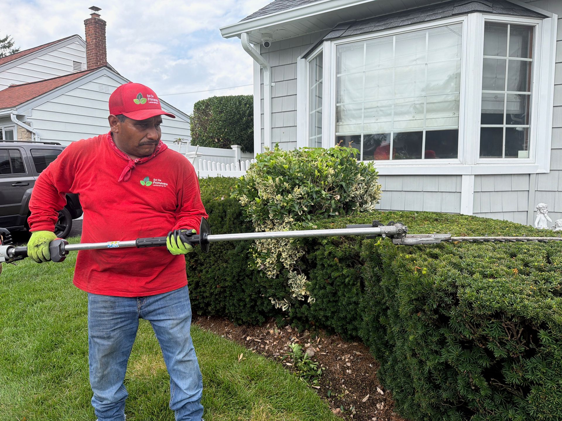 Worker in red uniform trimming a hedge with a long trimmer outside a house.