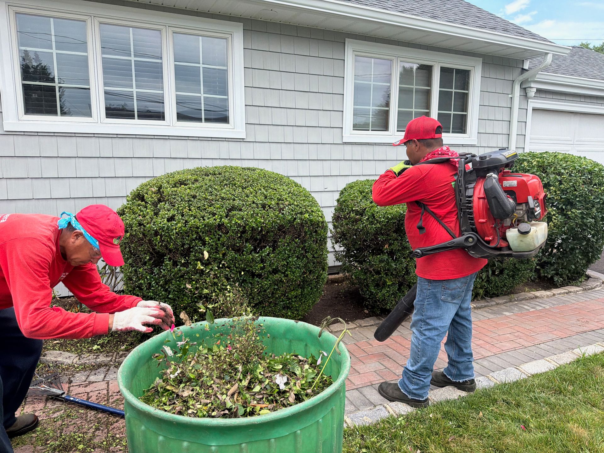 Two workers in red uniforms trimming shrubs beside a house with a leaf blower and yard waste bin.