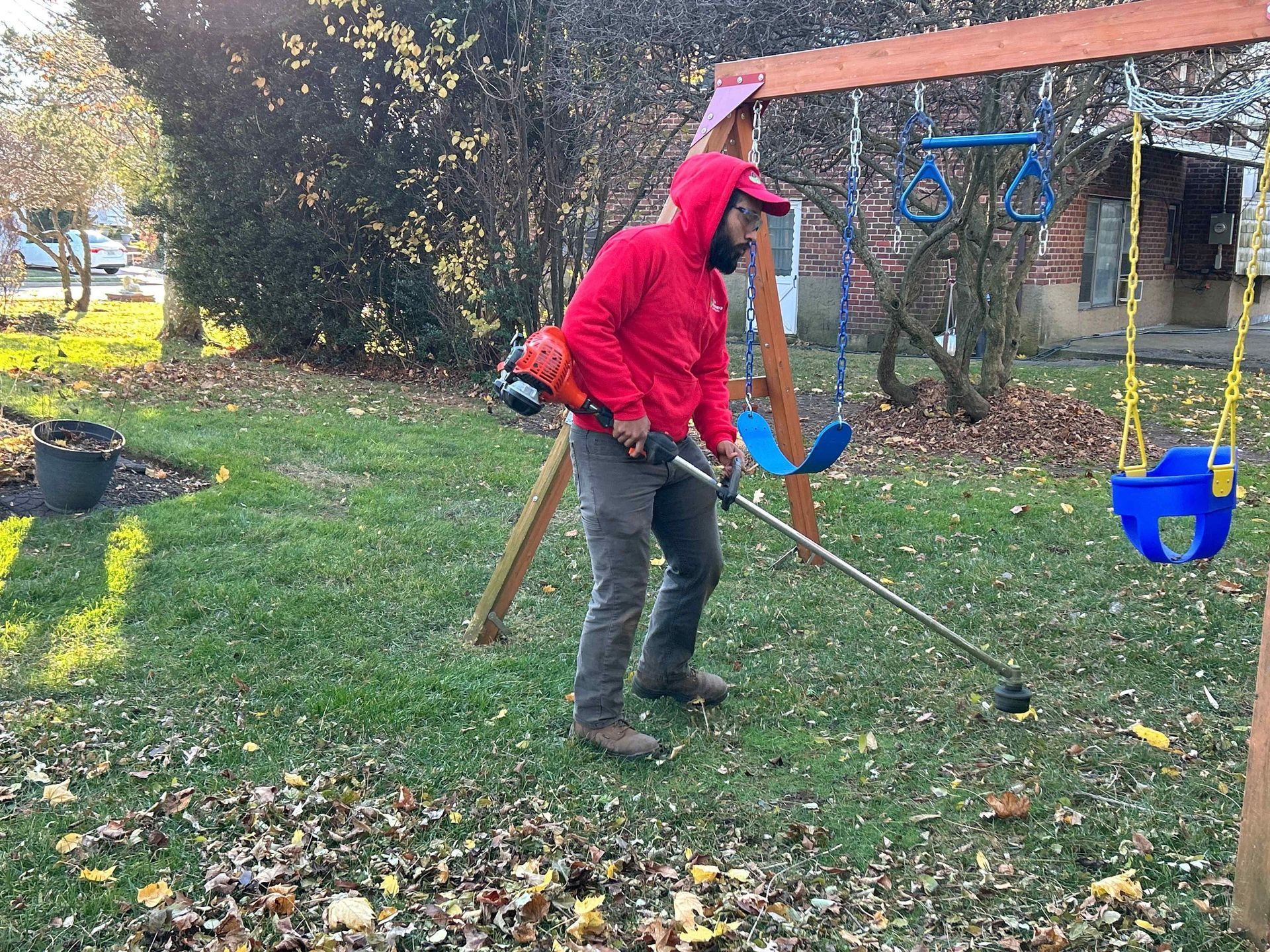 Person in a red hoodie using a string trimmer in a leafy backyard, near a blue swing set.