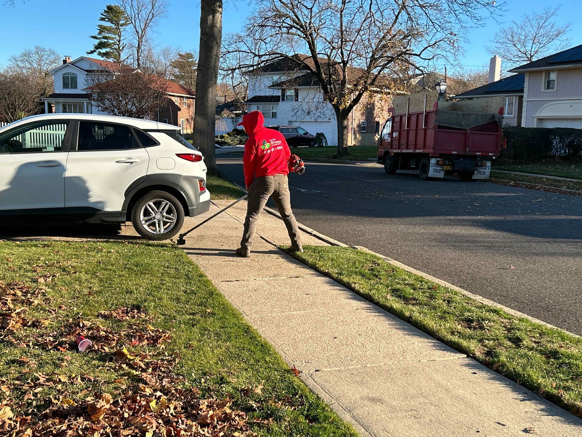 Person in a red hoodie raking leaves on a sidewalk in a suburban neighborhood