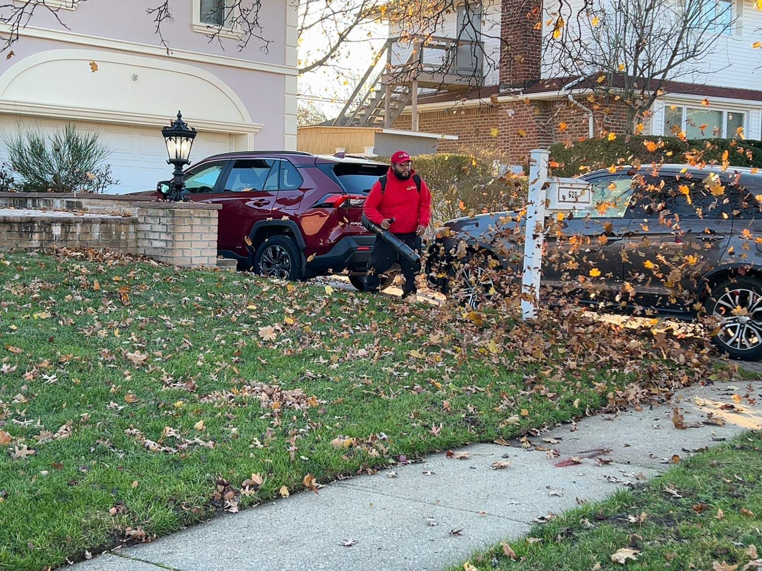 Person in red jacket crouches beside a red SUV in a leaf-covered suburban driveway.