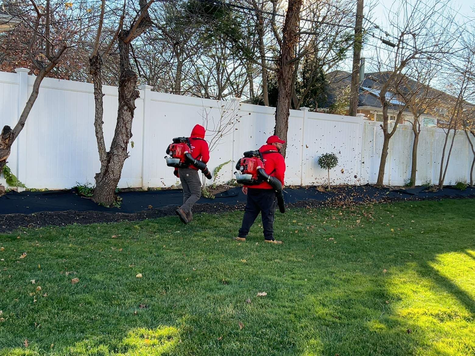 Two people in red jackets with backpacks walking near a white wall in a grassy yard
