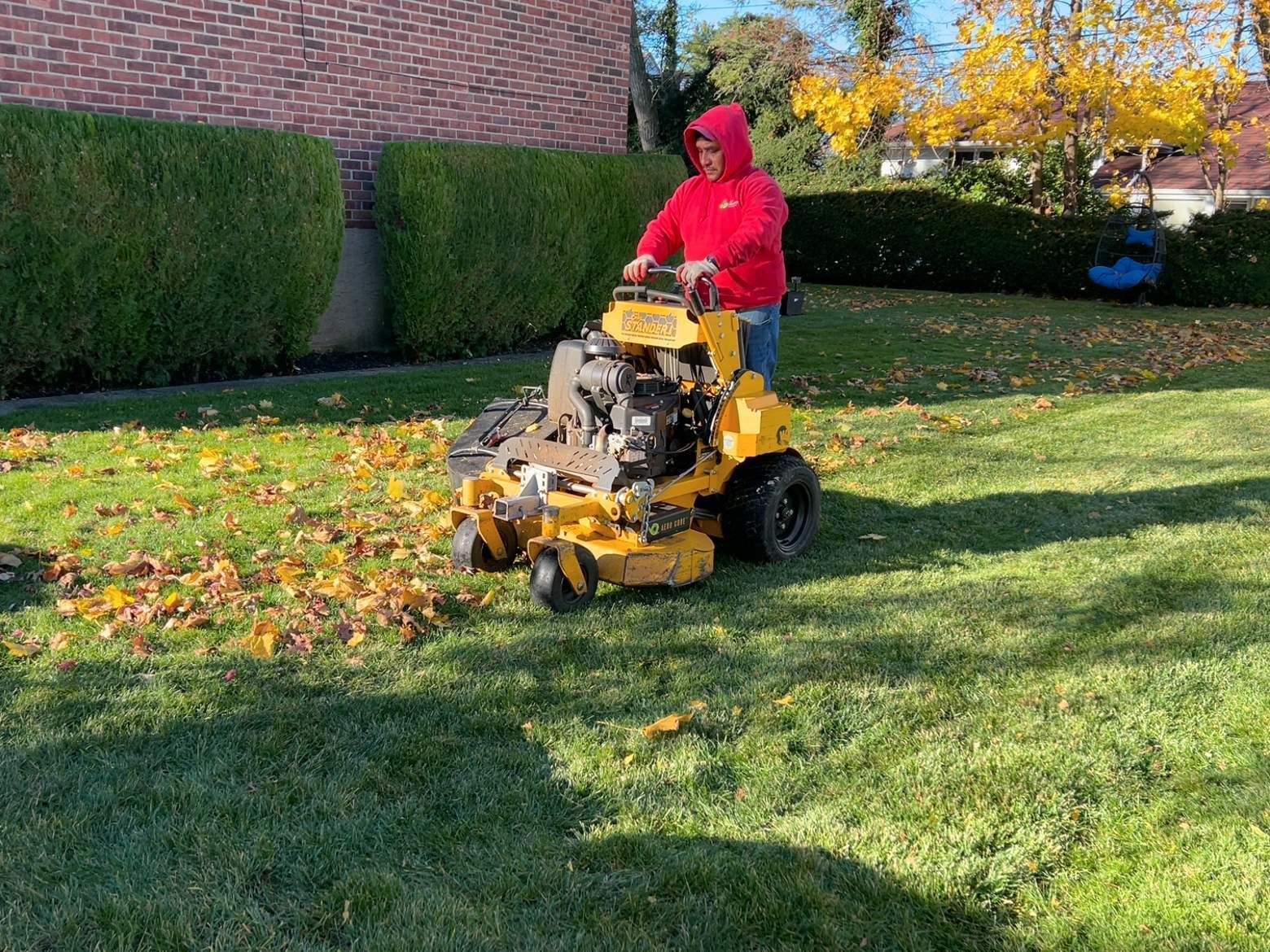 Person in a red hoodie mowing a leaf-covered lawn with a yellow riding mower.