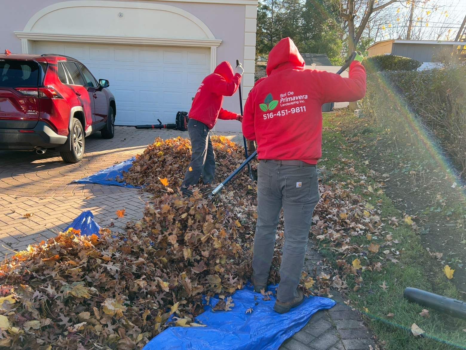Two workers in red hoodies rake autumn leaves beside a driveway and garage, with a red SUV nearby.