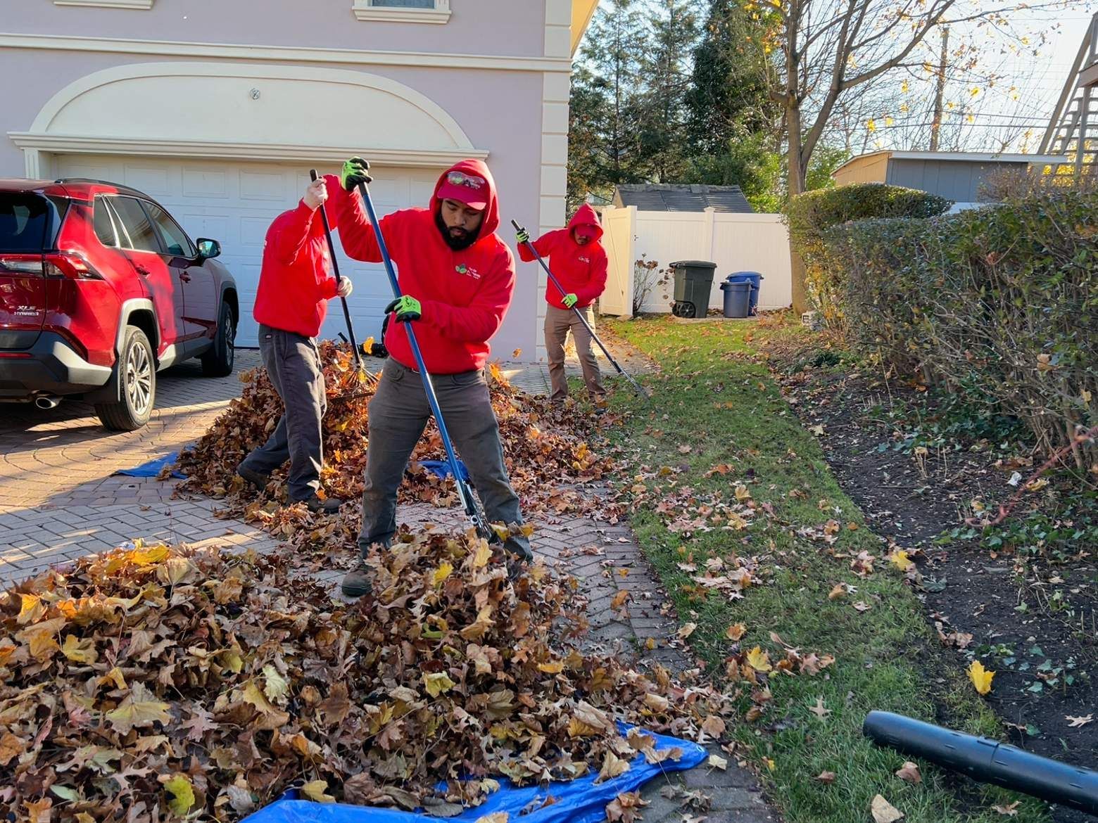 Workers in red jackets raking leaves along a driveway beside a house and parked cars