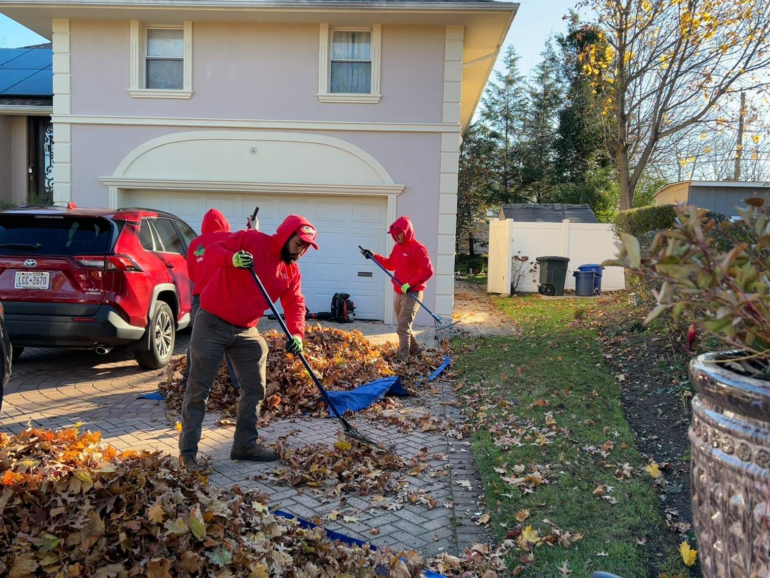 Workers in red jackets clearing leaves from a front yard beside a white two-story house.