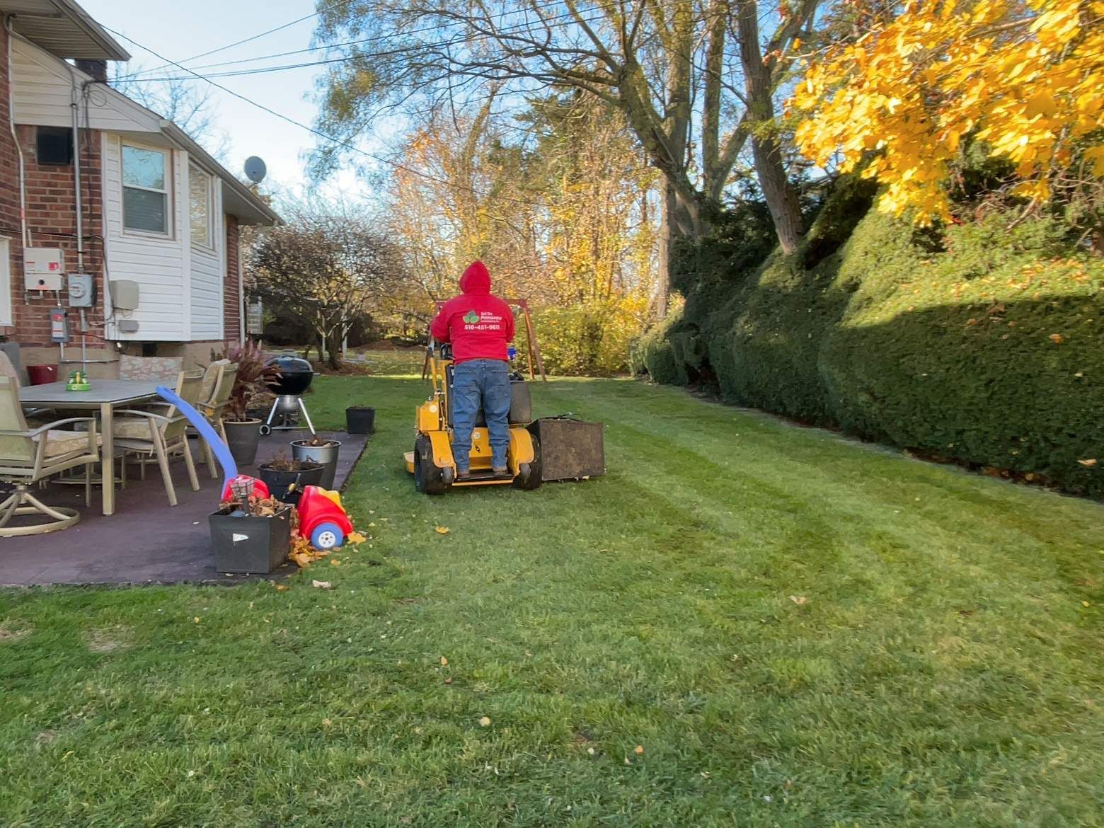 Person in red hoodie using a yellow lawn tractor in a grassy backyard with autumn trees.