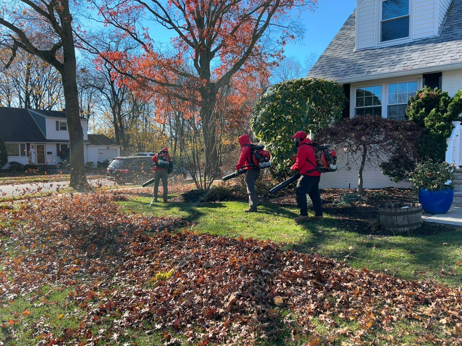 Workers in red jackets raking leaves in a suburban front yard beside a white house