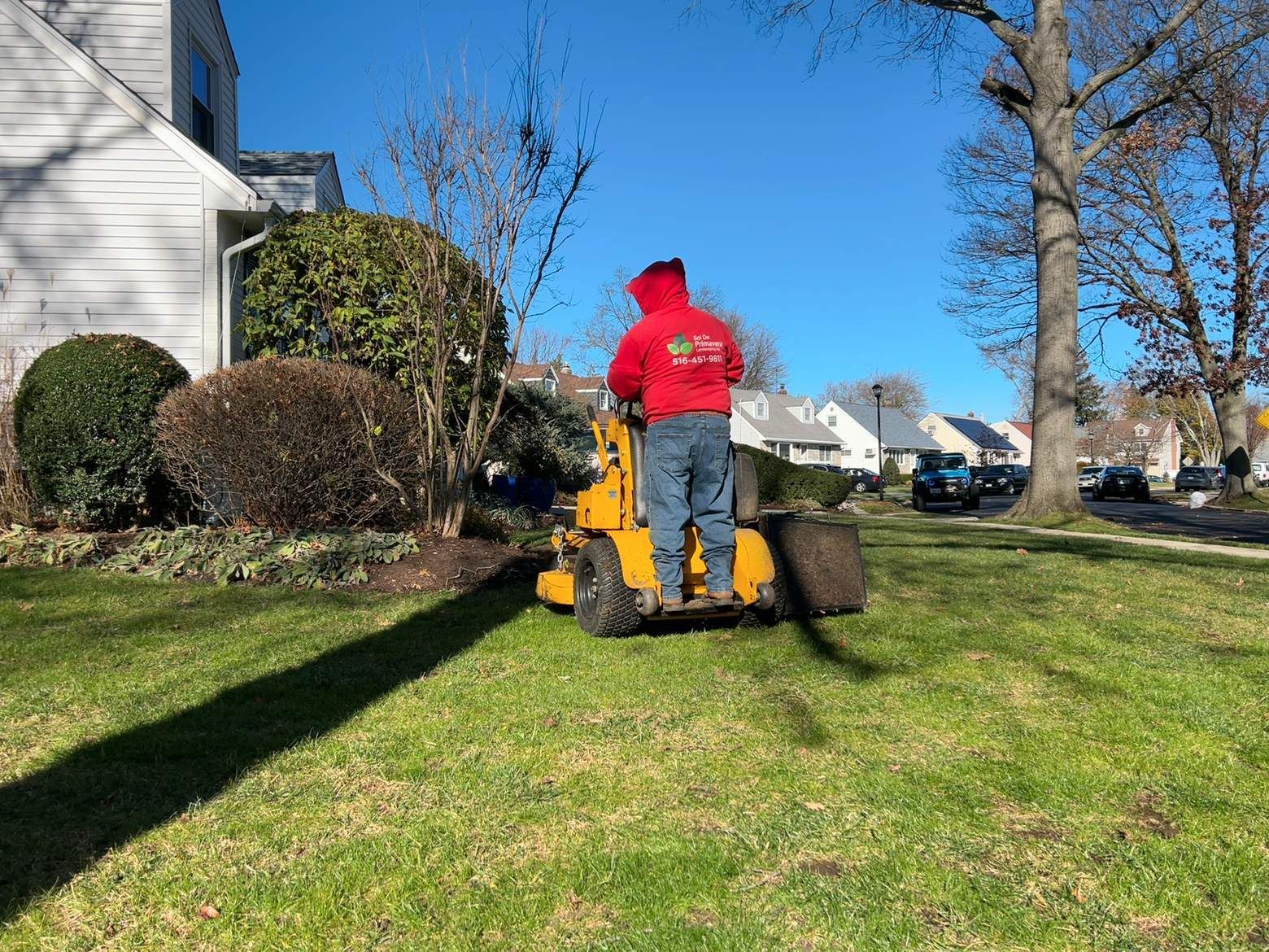 Person in a red jacket using a yellow lawn mower on a grassy suburban lawn.