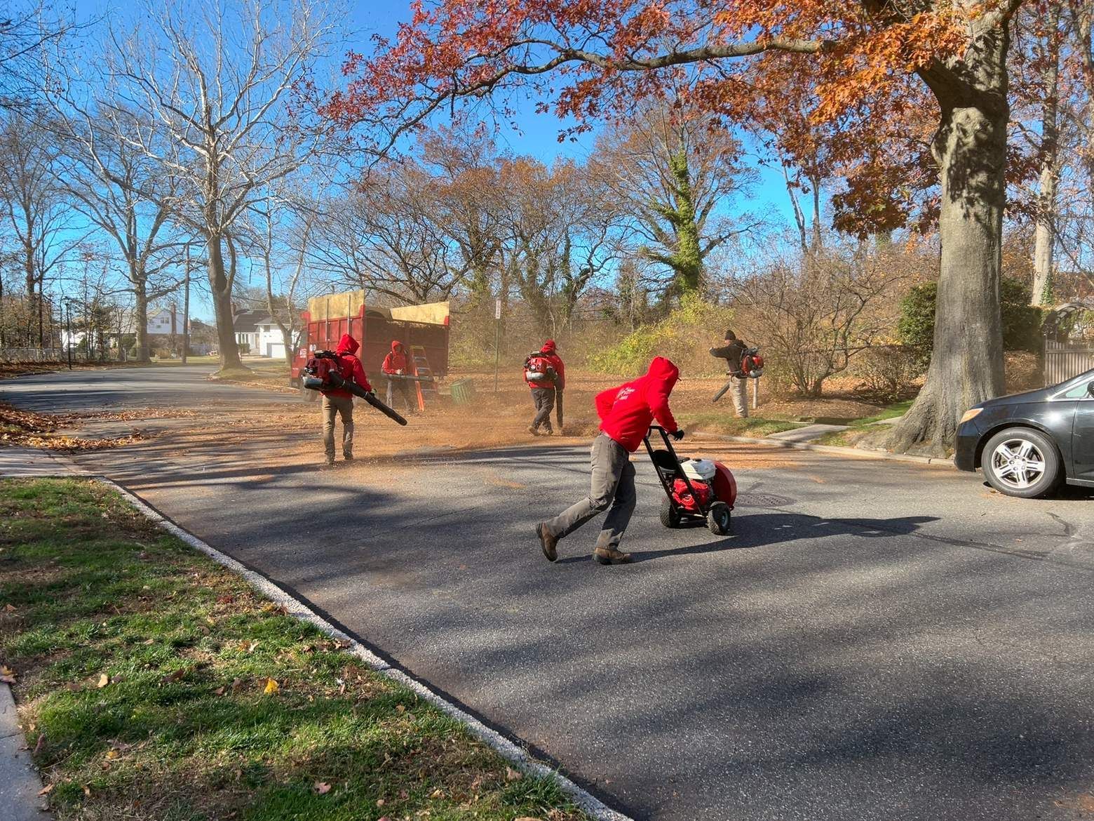 People in red jackets clear leaves on a suburban street in autumn.