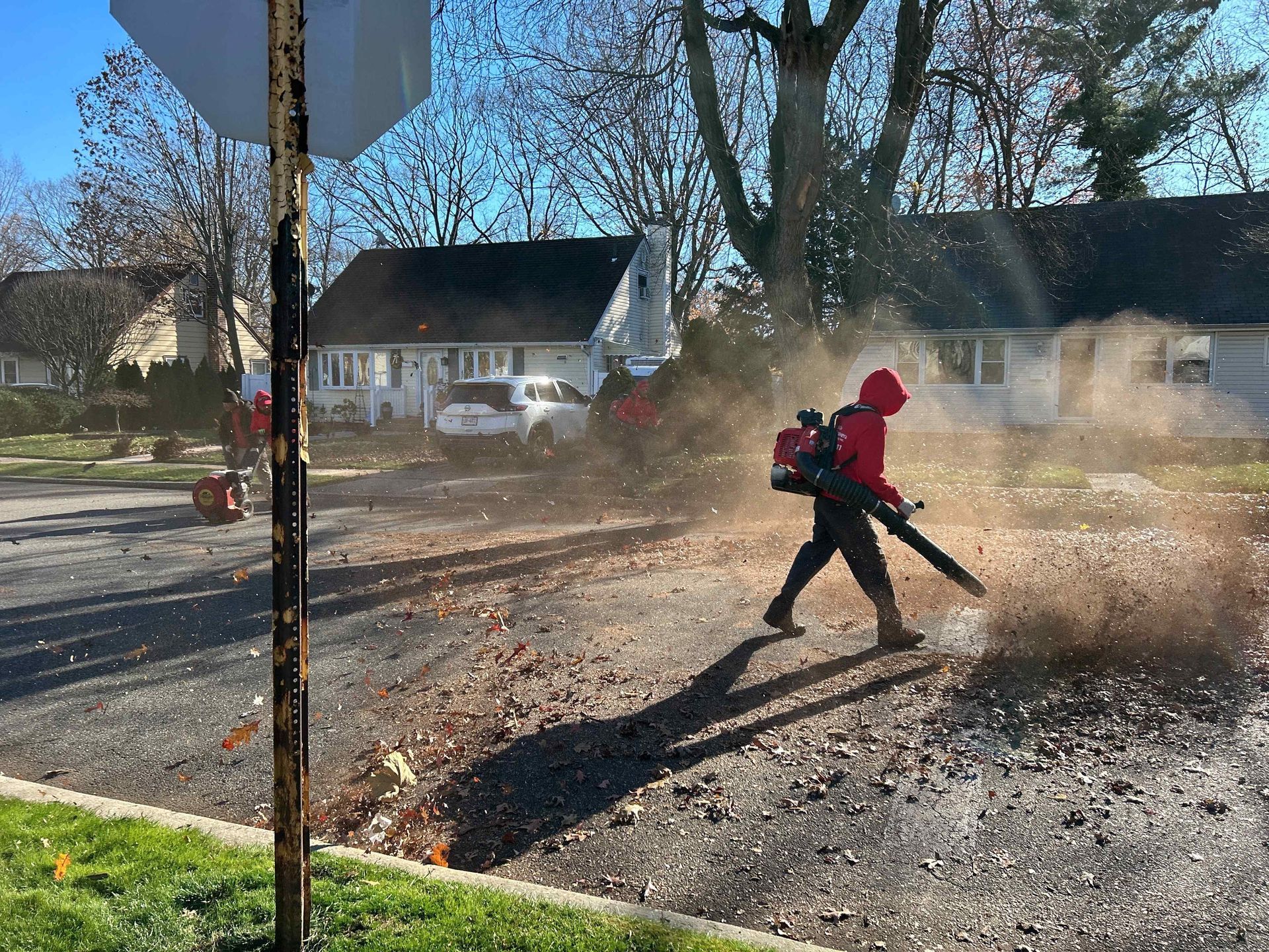 Worker in red using a leaf blower on a dusty street near houses and bare trees