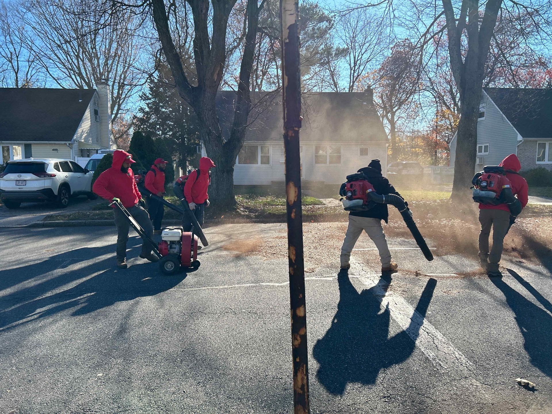 People in red jackets raking leaves in a sunny residential driveway