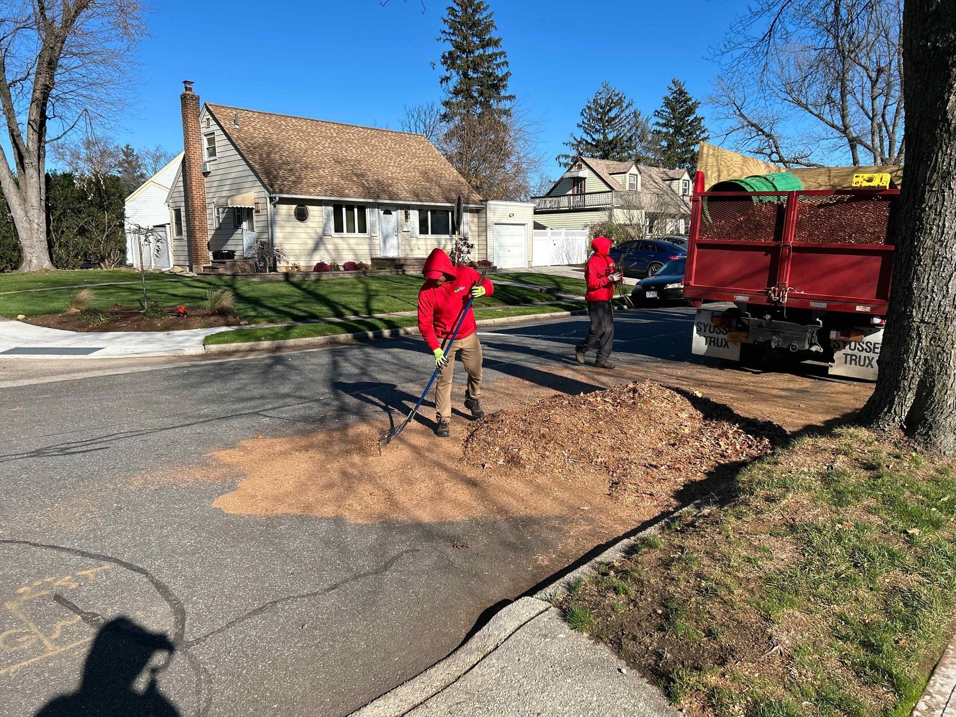 Workers clearing fallen leaves on a suburban street near a red truck and houses