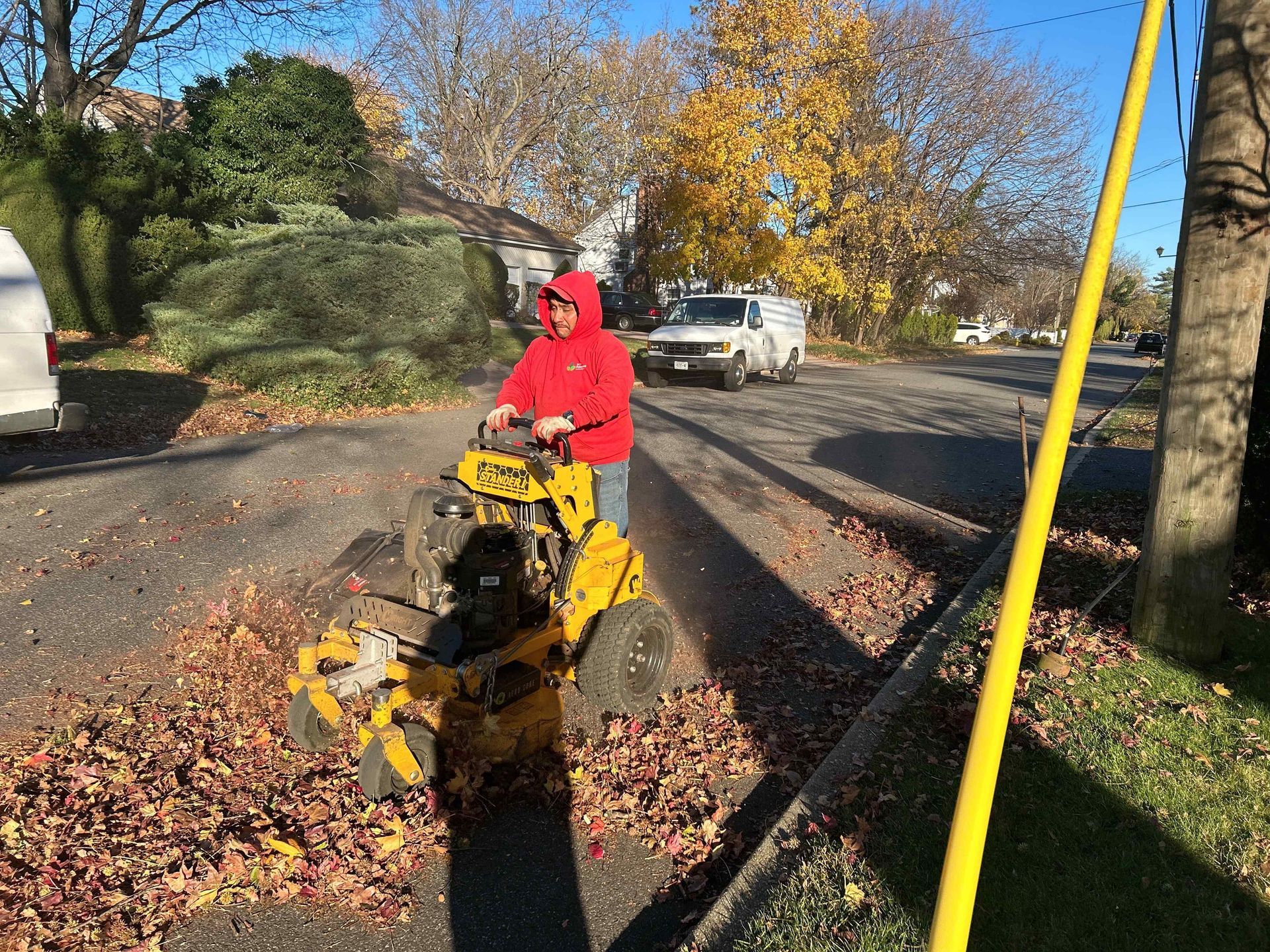 Person in red jacket using yellow leaf blower on a leaf-covered street, with parked car and autumn trees.