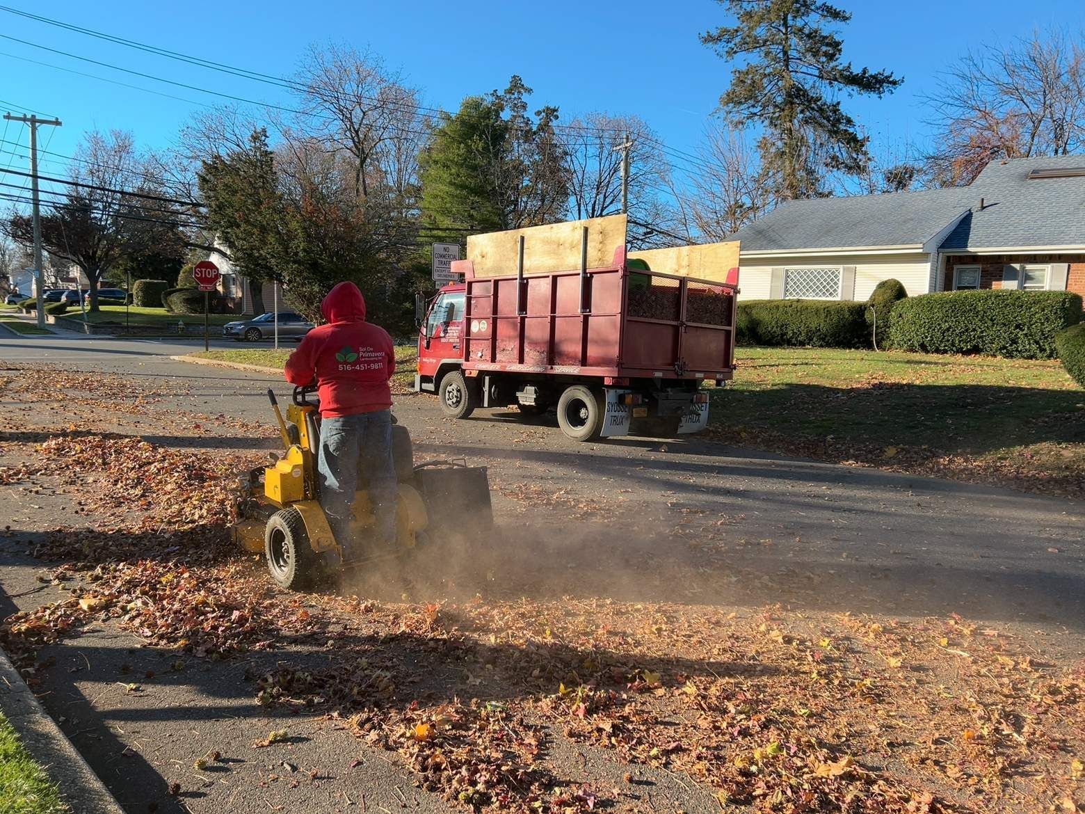 Worker in red hoodie using a leaf blower beside a red dump trailer on a leafy residential street.