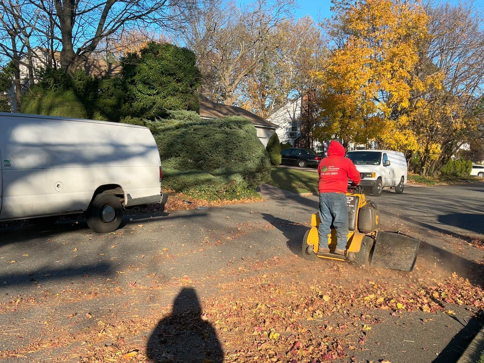 Worker in red jacket using a wheelbarrow on a leaf-covered driveway beside parked vans and autumn trees