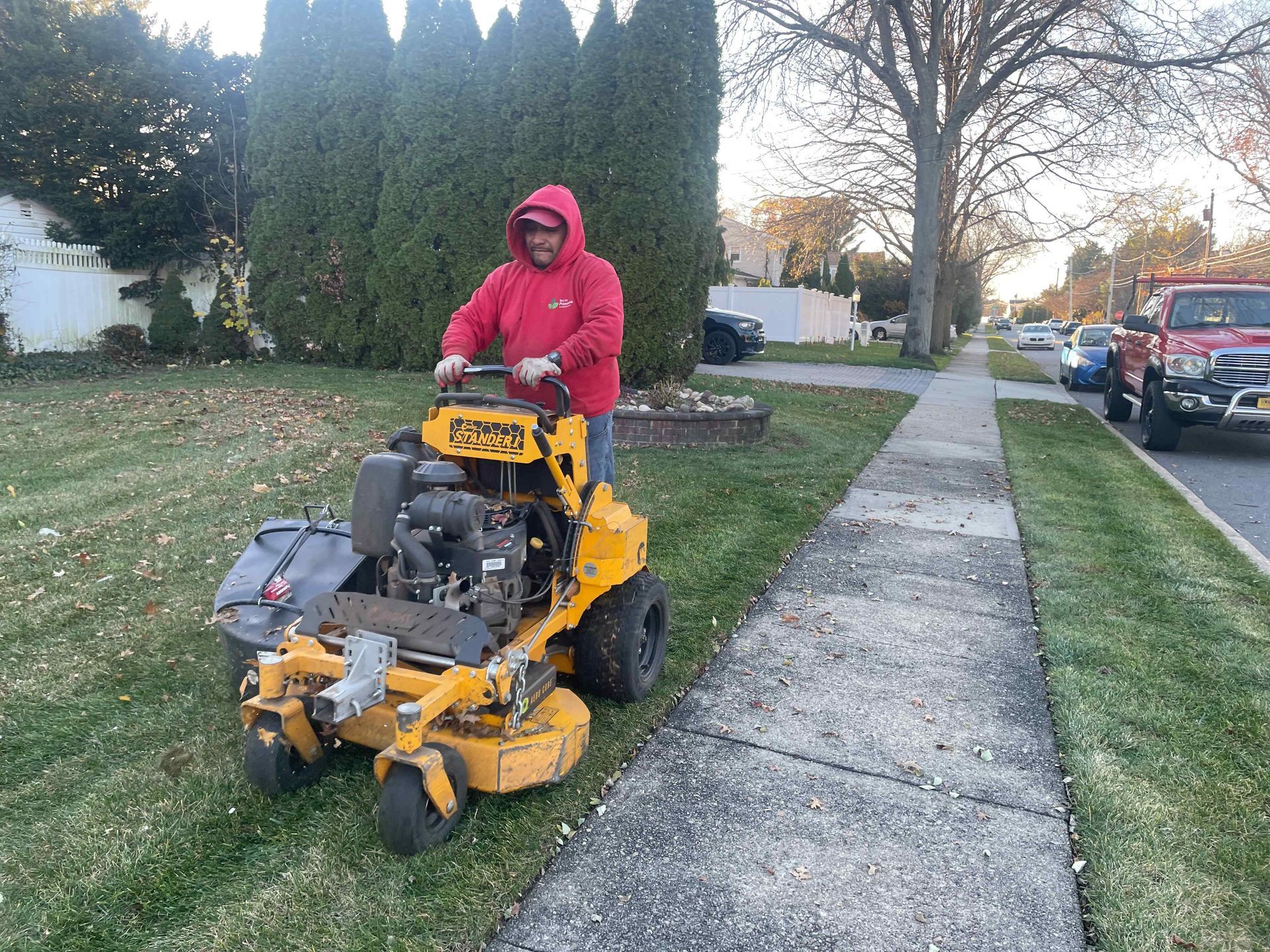Person in a red hoodie mowing a grassy sidewalk strip with a yellow riding mower on a suburban street.