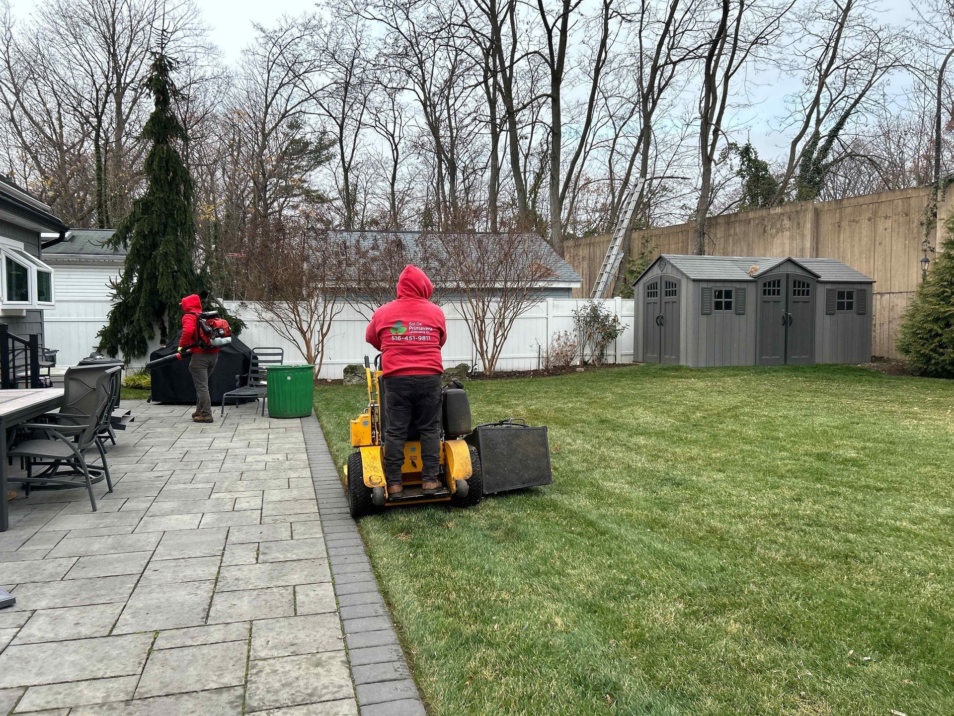 Two people in red jackets using lawn equipment on a backyard lawn near a patio and storage sheds