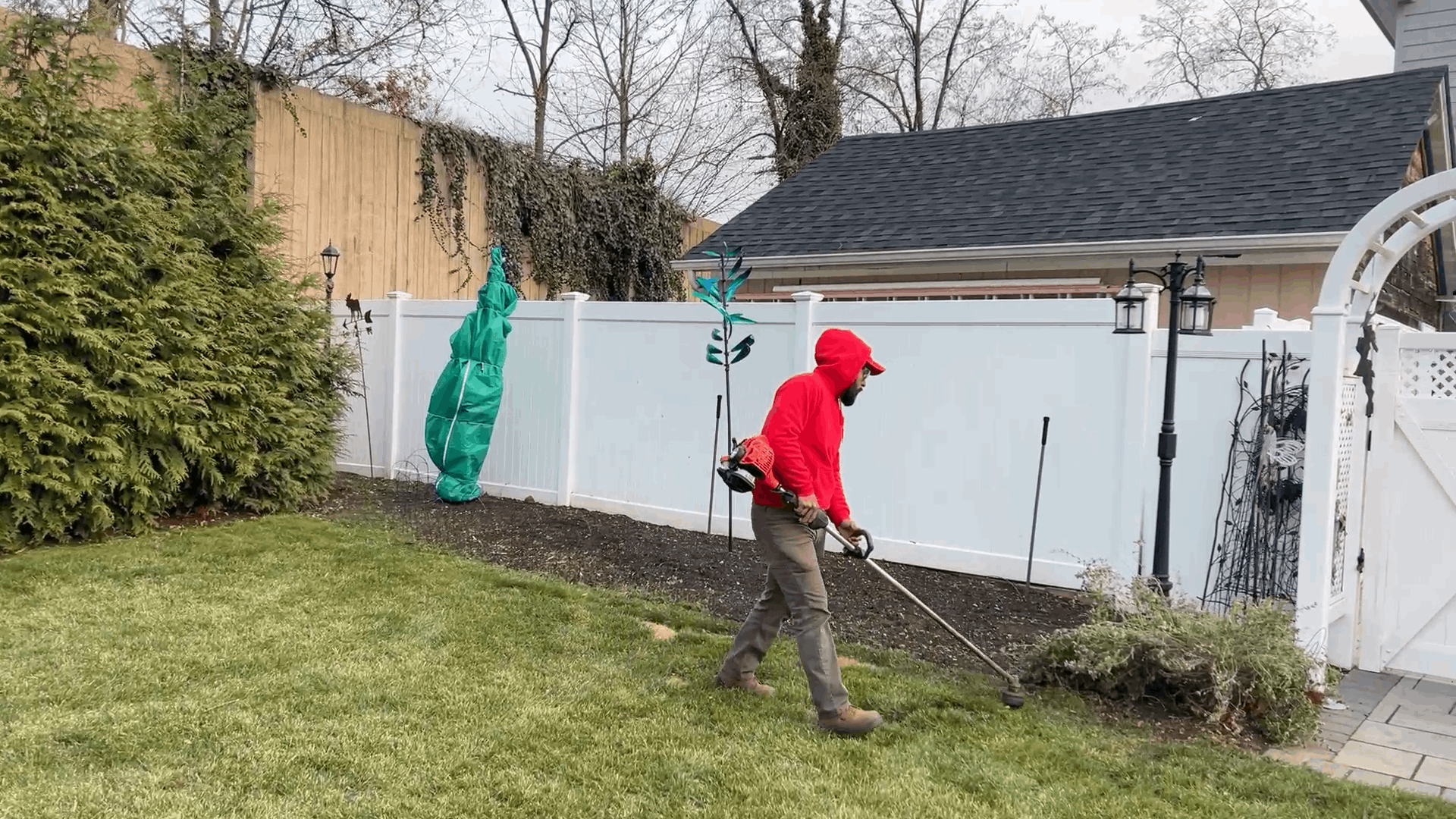 Person in red hoodie and green pants mowing a backyard lawn near a white fence and shed