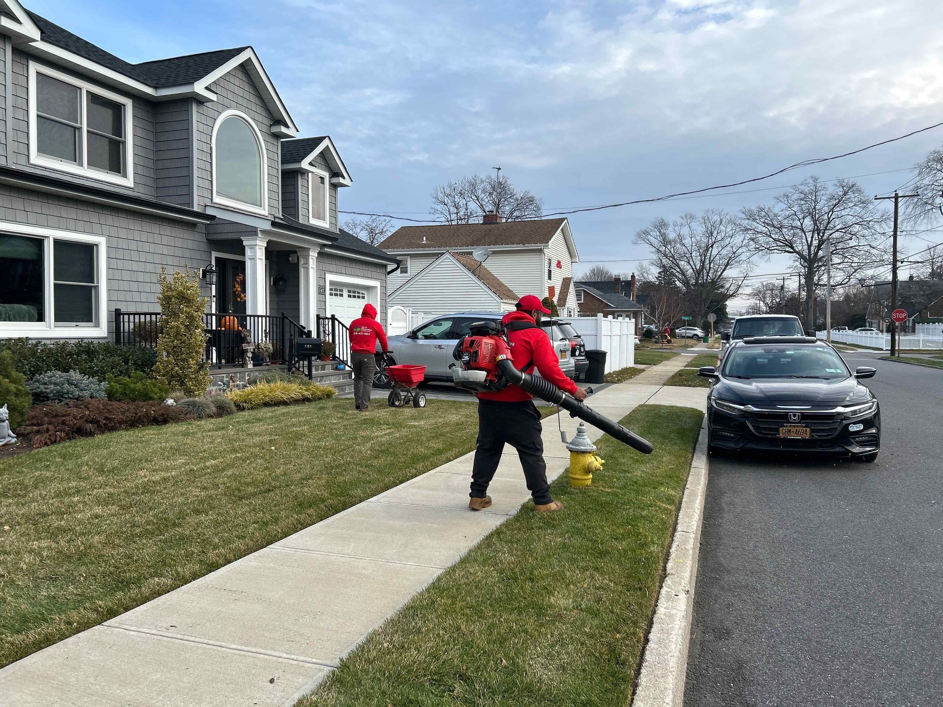 Workers in red jackets using a leaf blower on a suburban sidewalk beside a parked car
