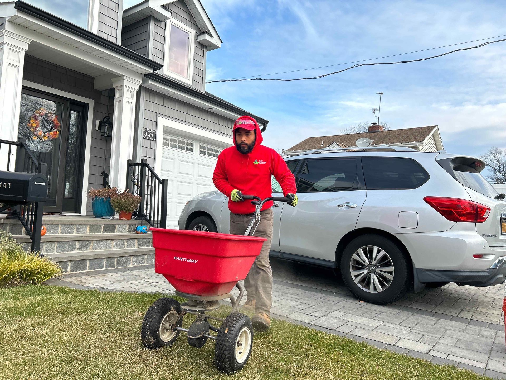 Person in a red jacket pushing a red spreader on a driveway beside a silver SUV and house
