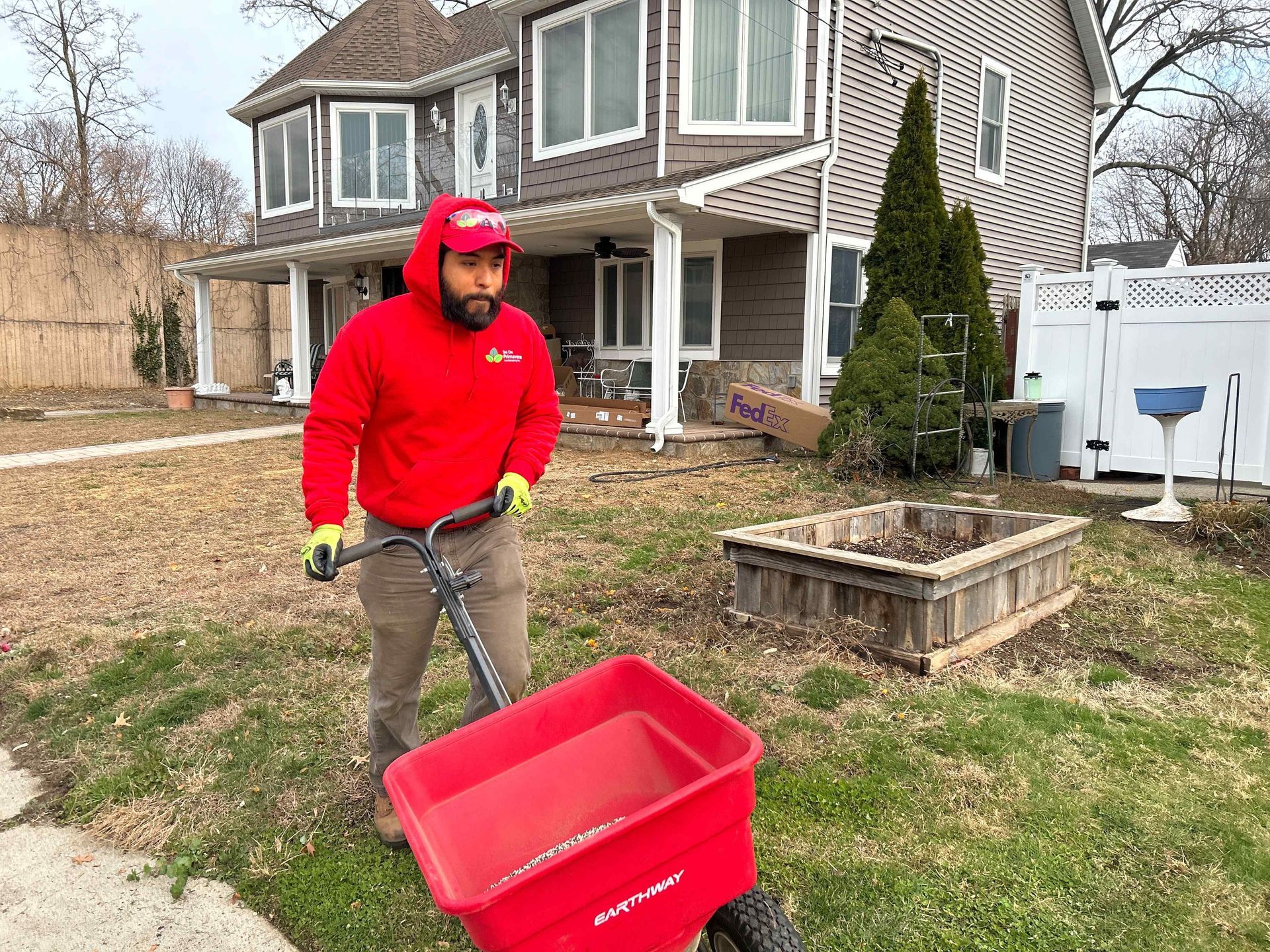 Person in a red hoodie pushing a red yard cart in a suburban yard beside a house.