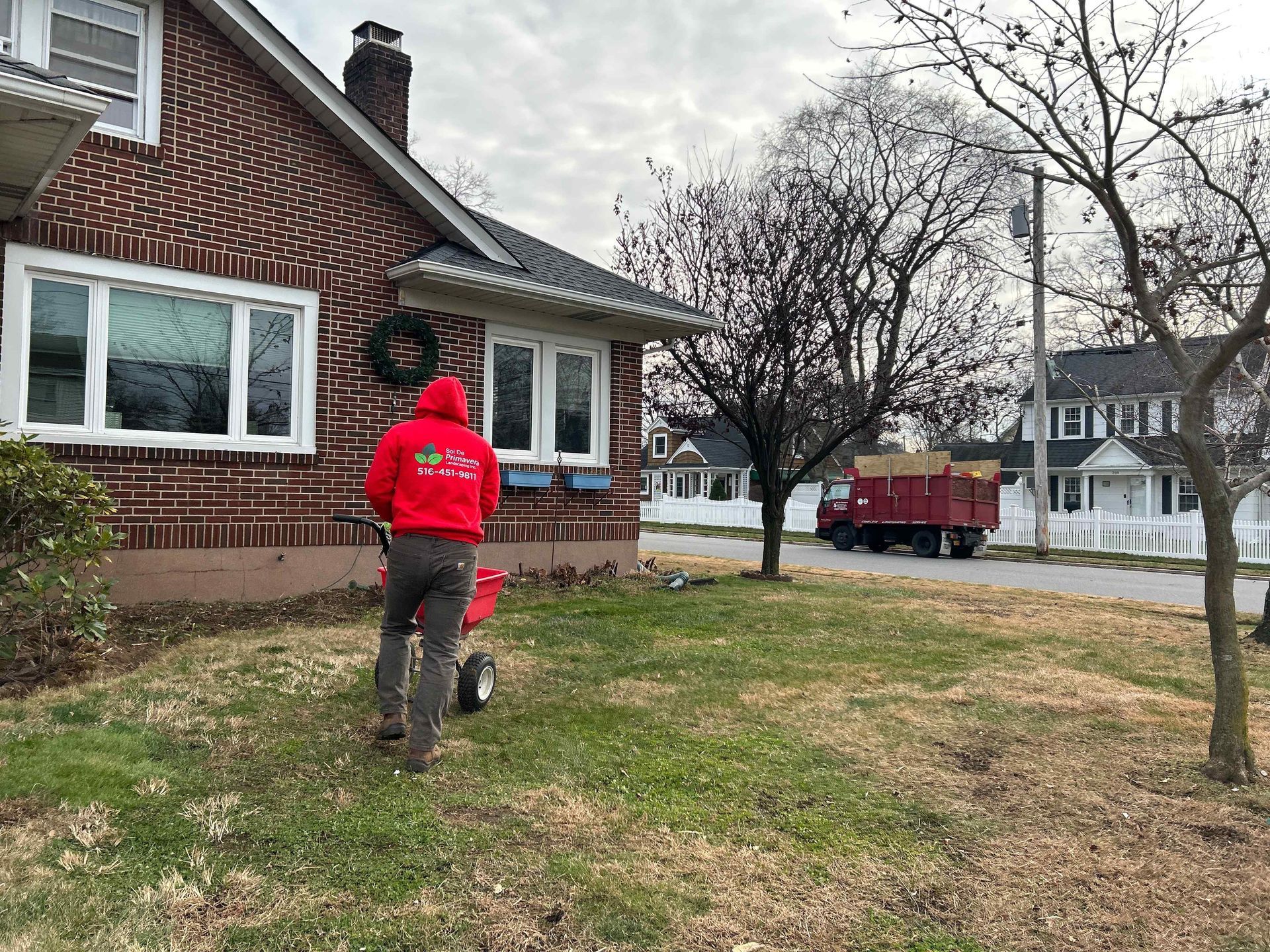 Person in a red hoodie walking a small dog in a yard beside a brick house on an overcast day