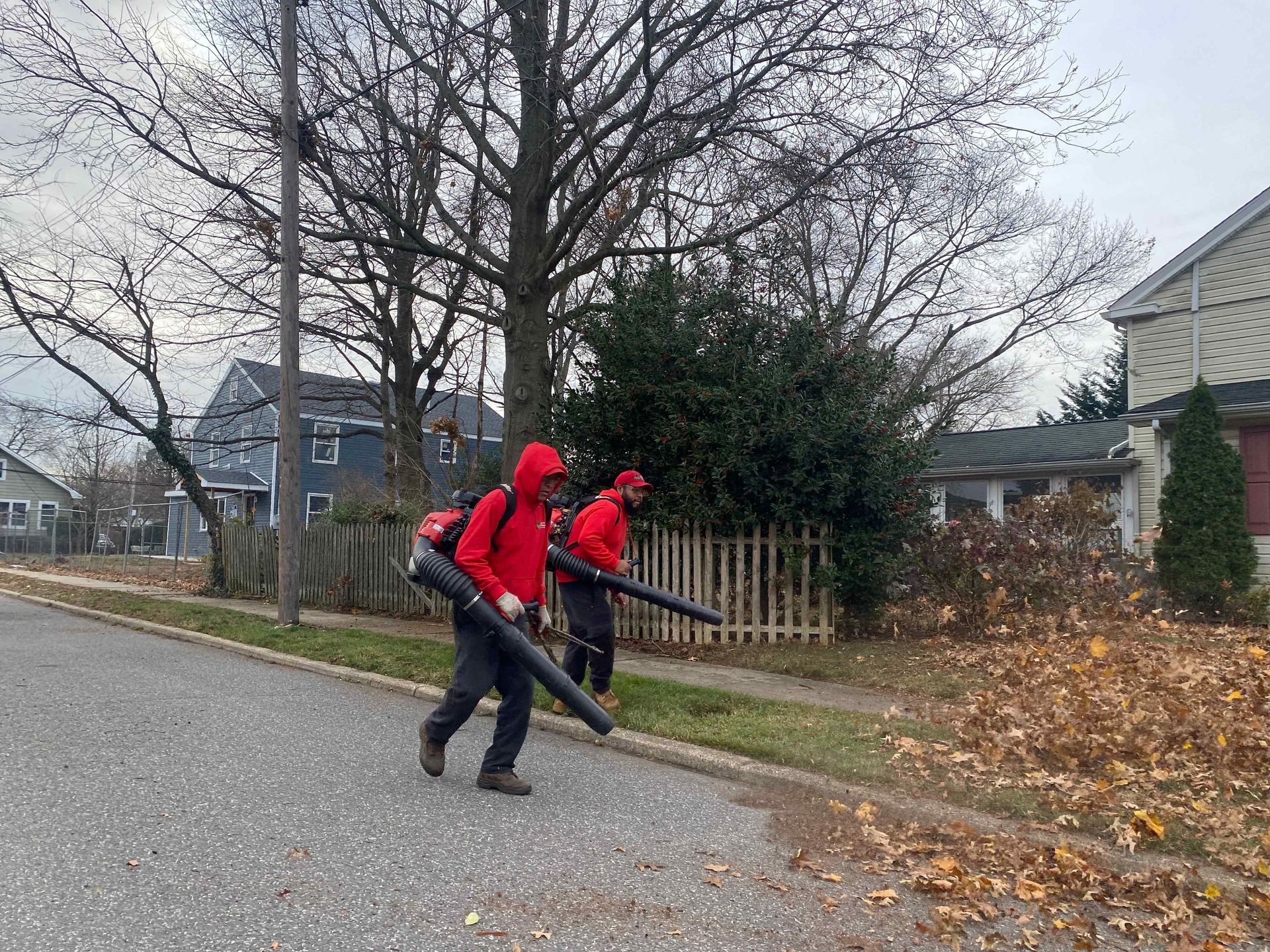 Two workers in red jackets and backpacks use leaf blowers along a suburban street