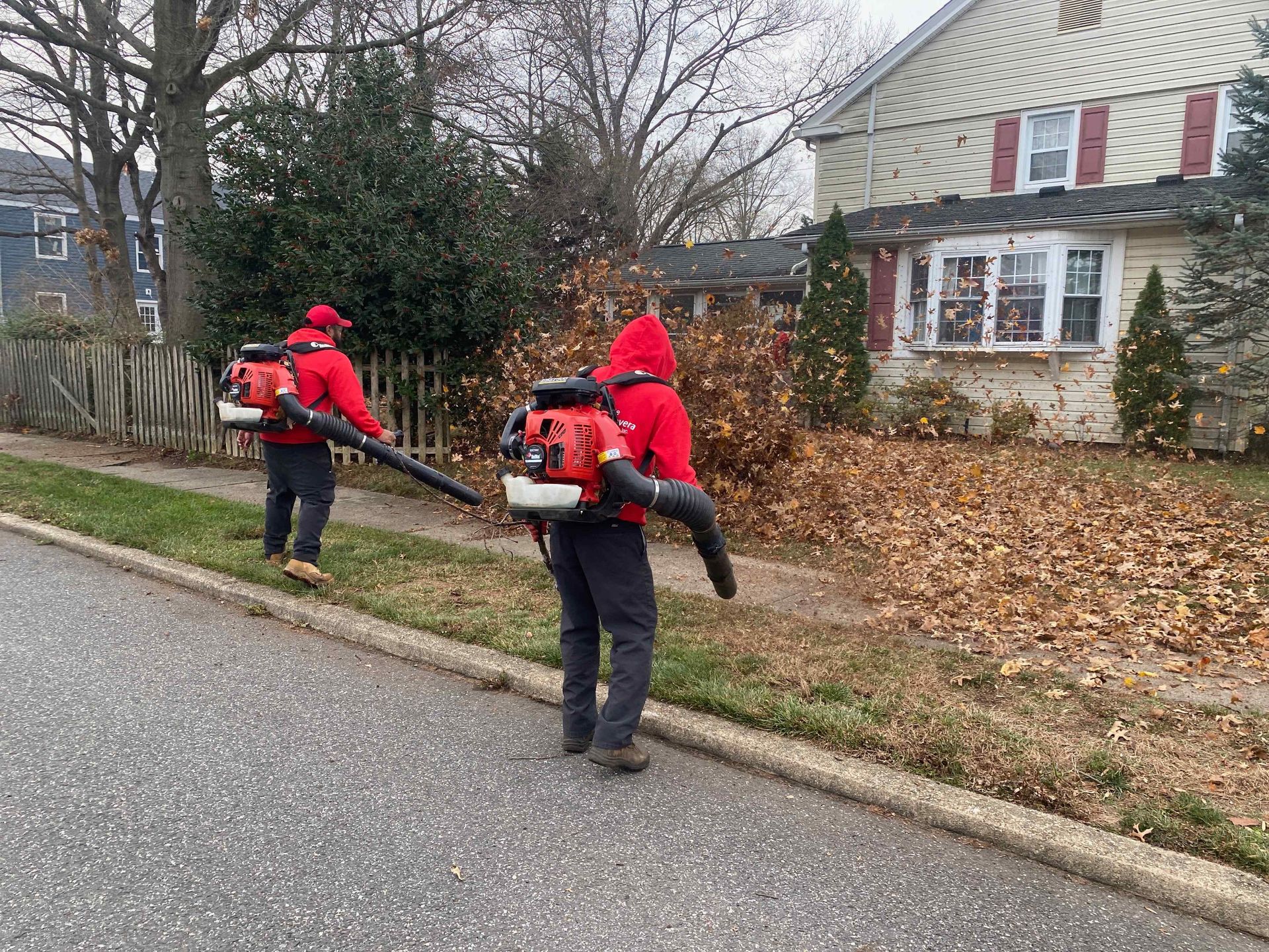 Two workers in red jackets use leaf blowers along a suburban roadside beside a house.