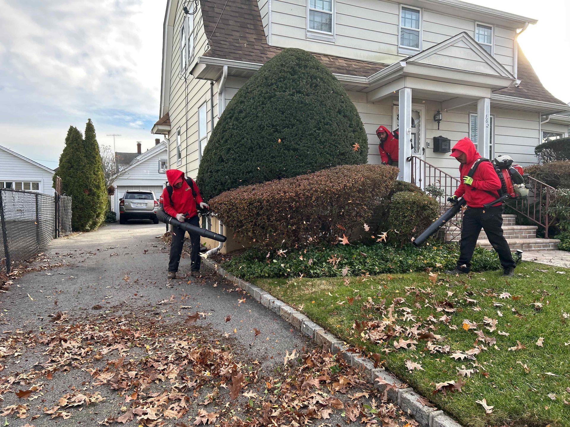 Two landscapers in red jackets blow leaves along a driveway beside a house and trimmed shrubs.