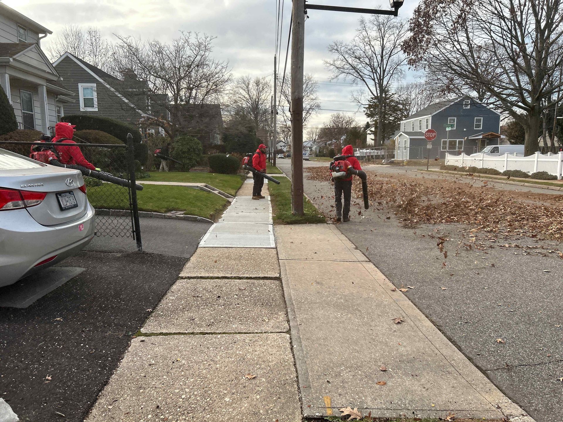 Three workers in red jackets clearing leaves along a suburban sidewalk near parked cars