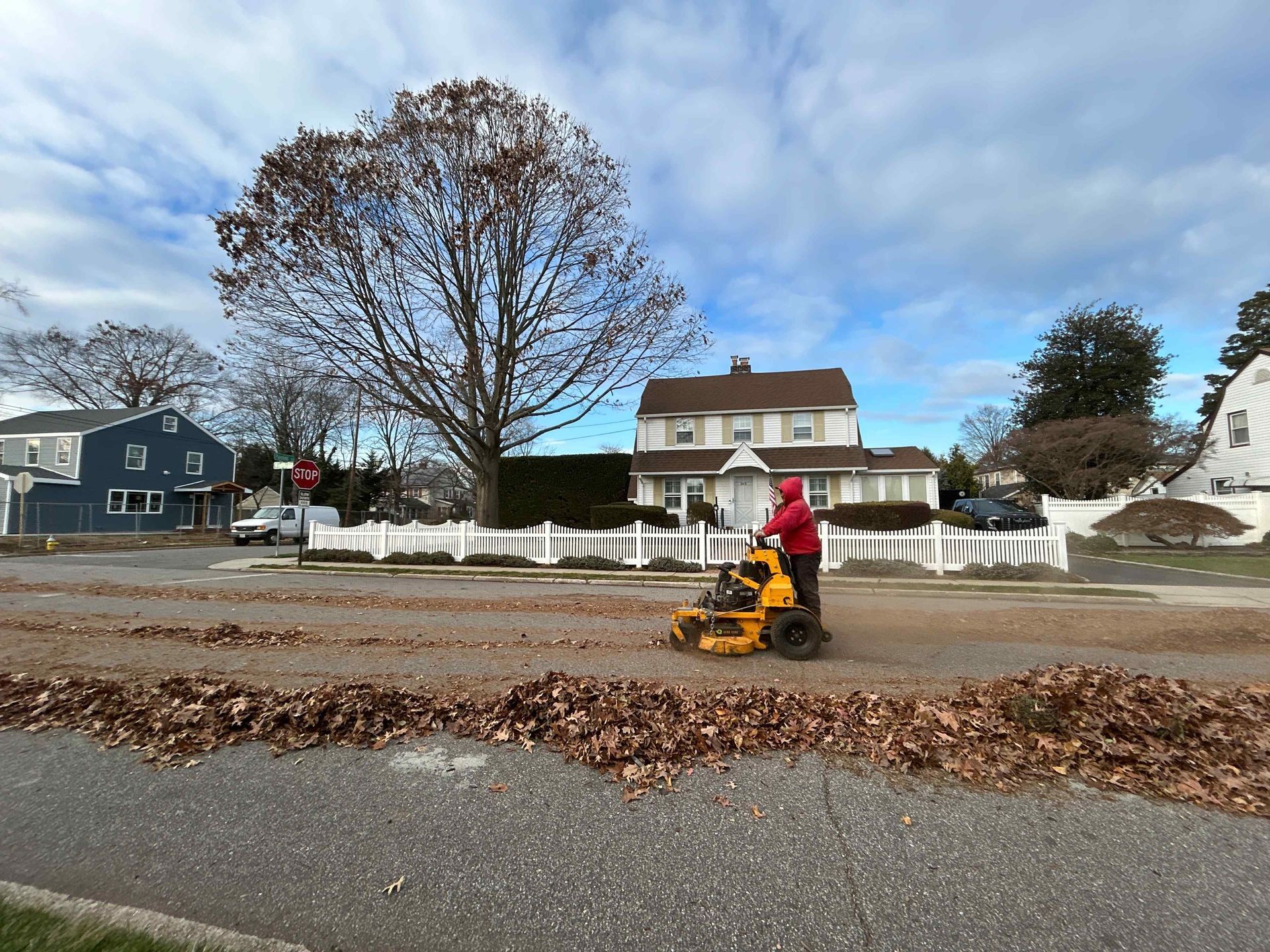 Person in red using a yellow leaf blower on a leaf-covered suburban street in front of houses