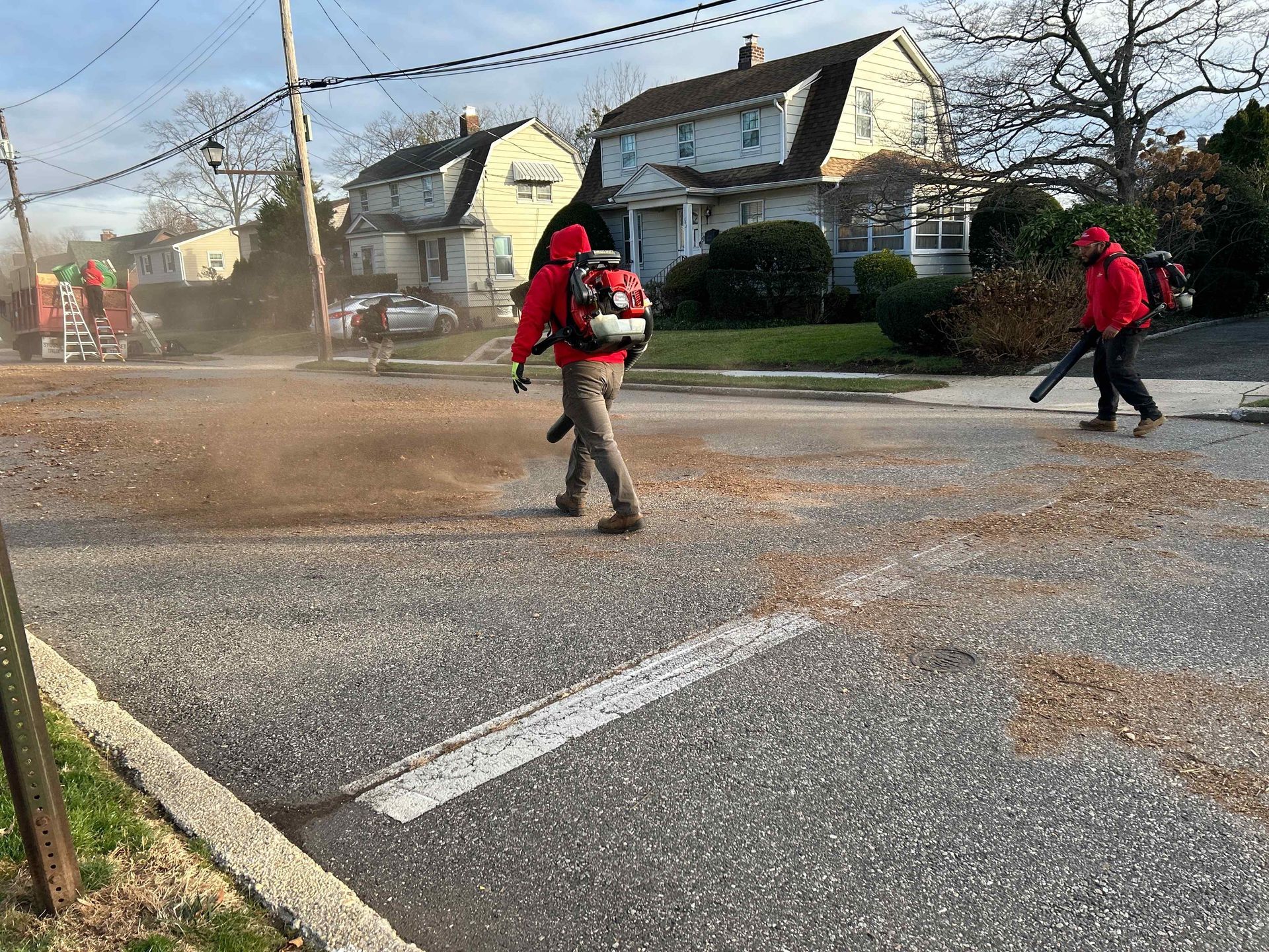 Two people in red jackets crossing a dusty street in front of houses and a road crew truck.