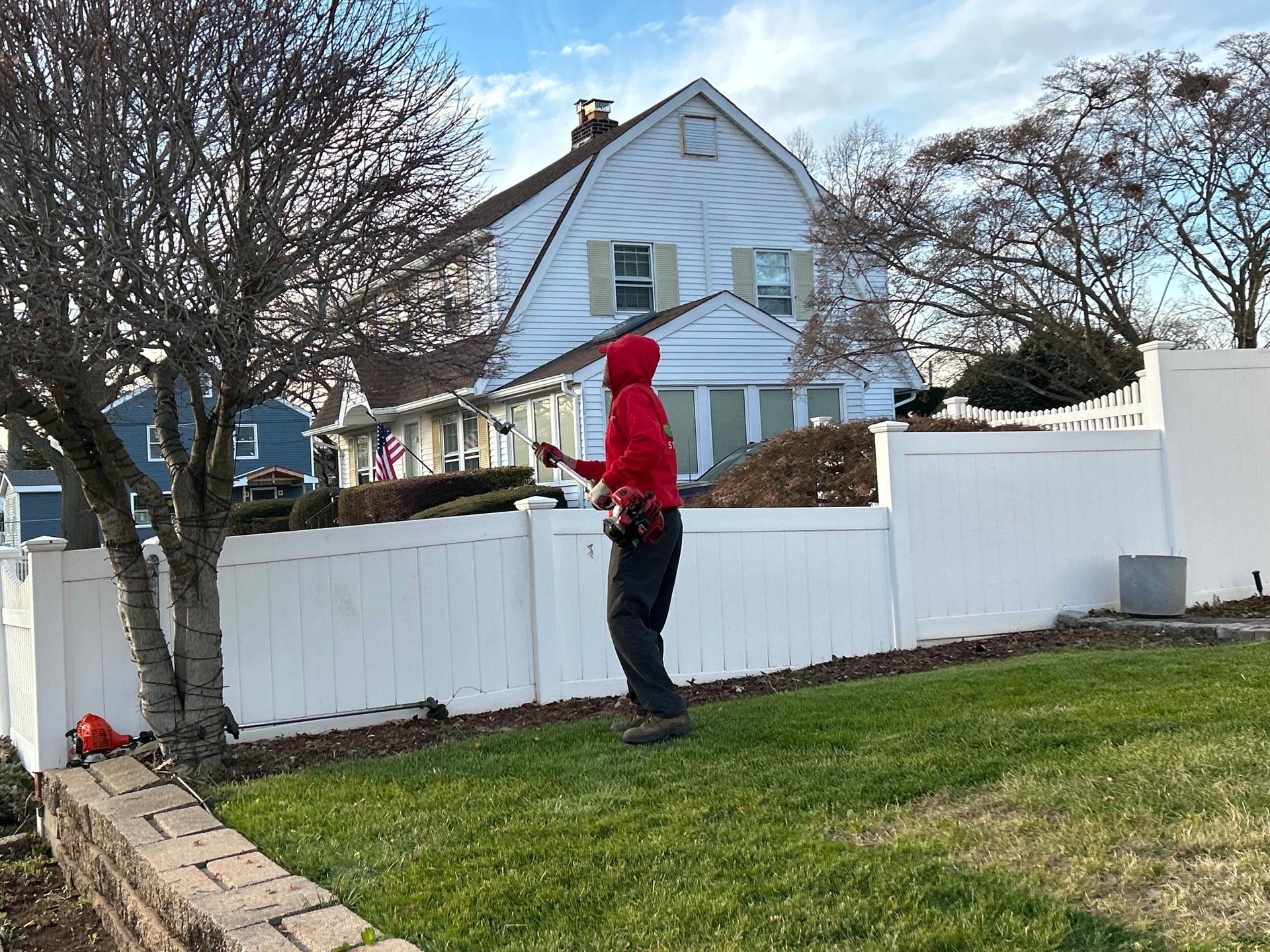 Person in red jacket standing by a white fence in a suburban yard with a blue house behind.