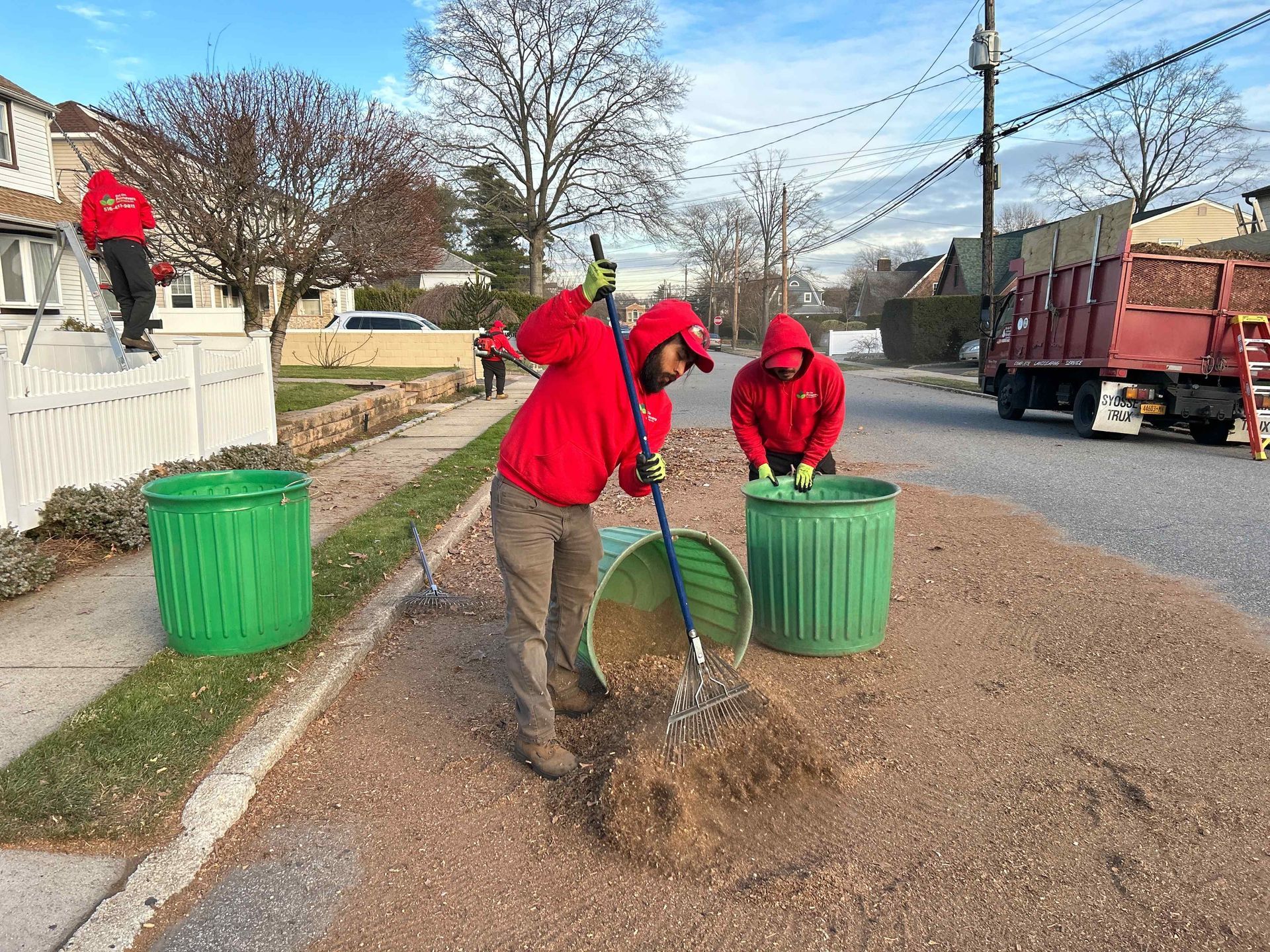 Workers in red hoodies cleaning leaves along a roadside, with green bins and a truck nearby.