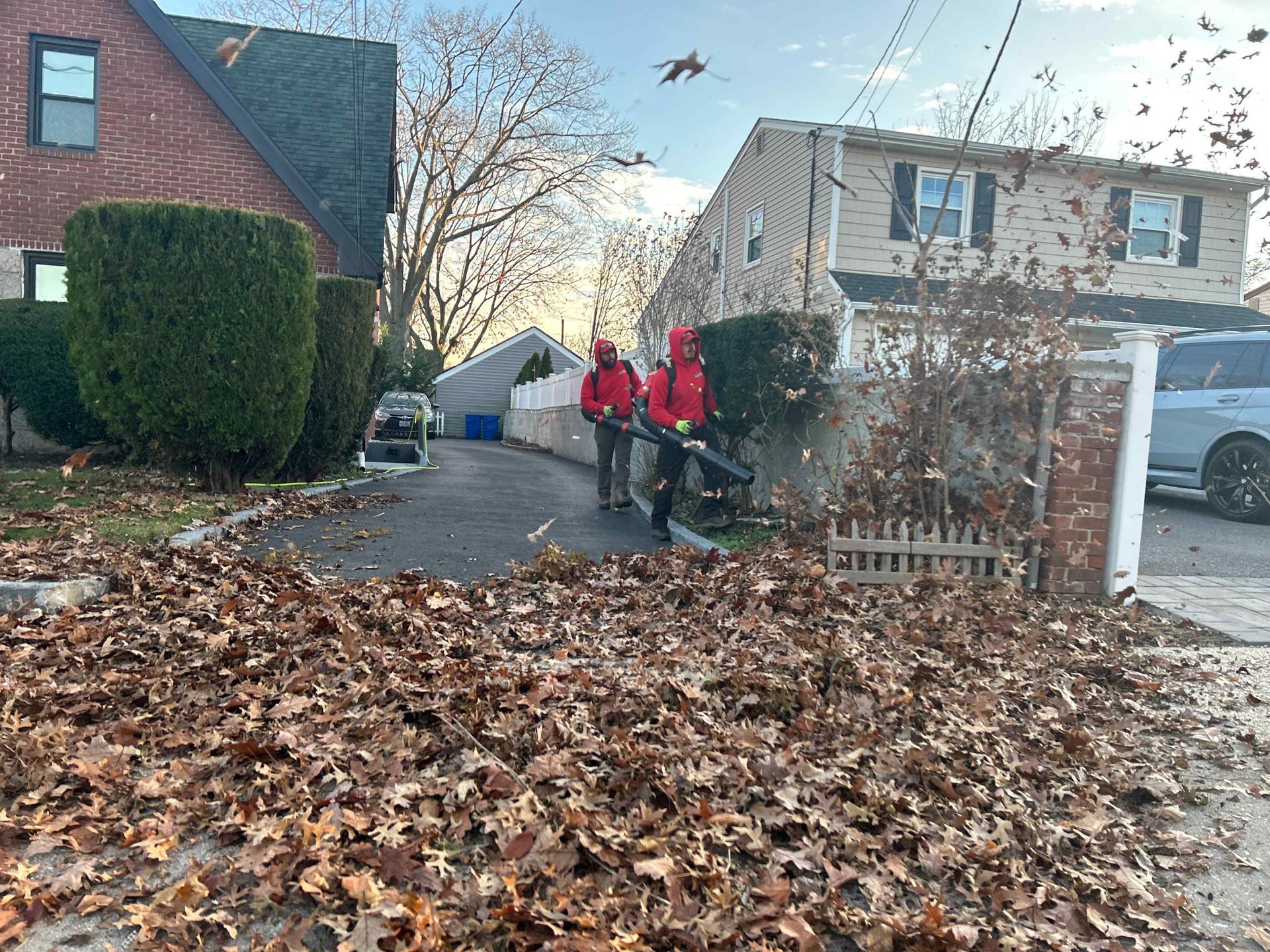 Three people in red jackets walking down a leaf-covered driveway beside suburban houses