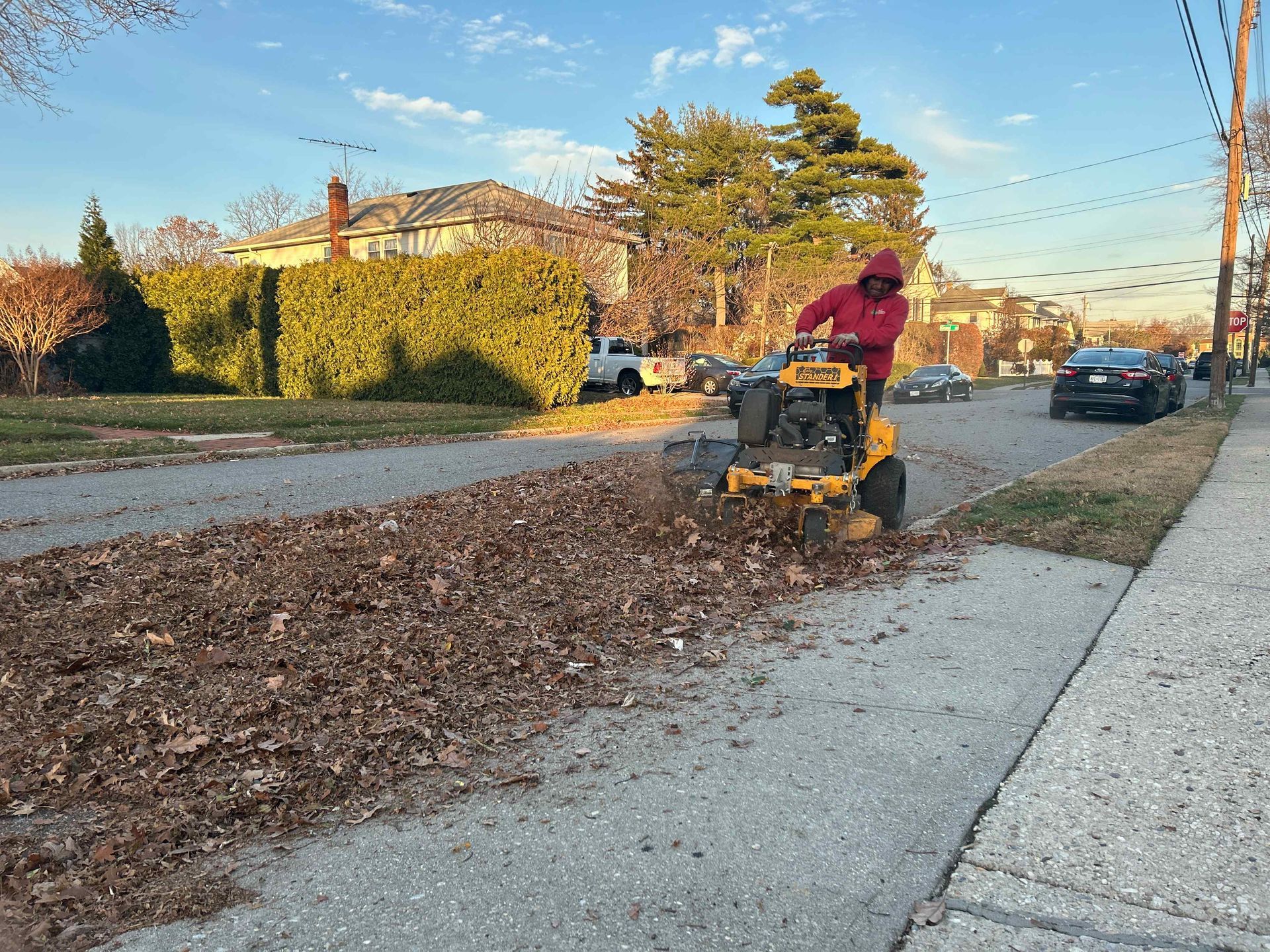 Worker using a leaf blower to clear fallen leaves from a suburban sidewalk at sunset