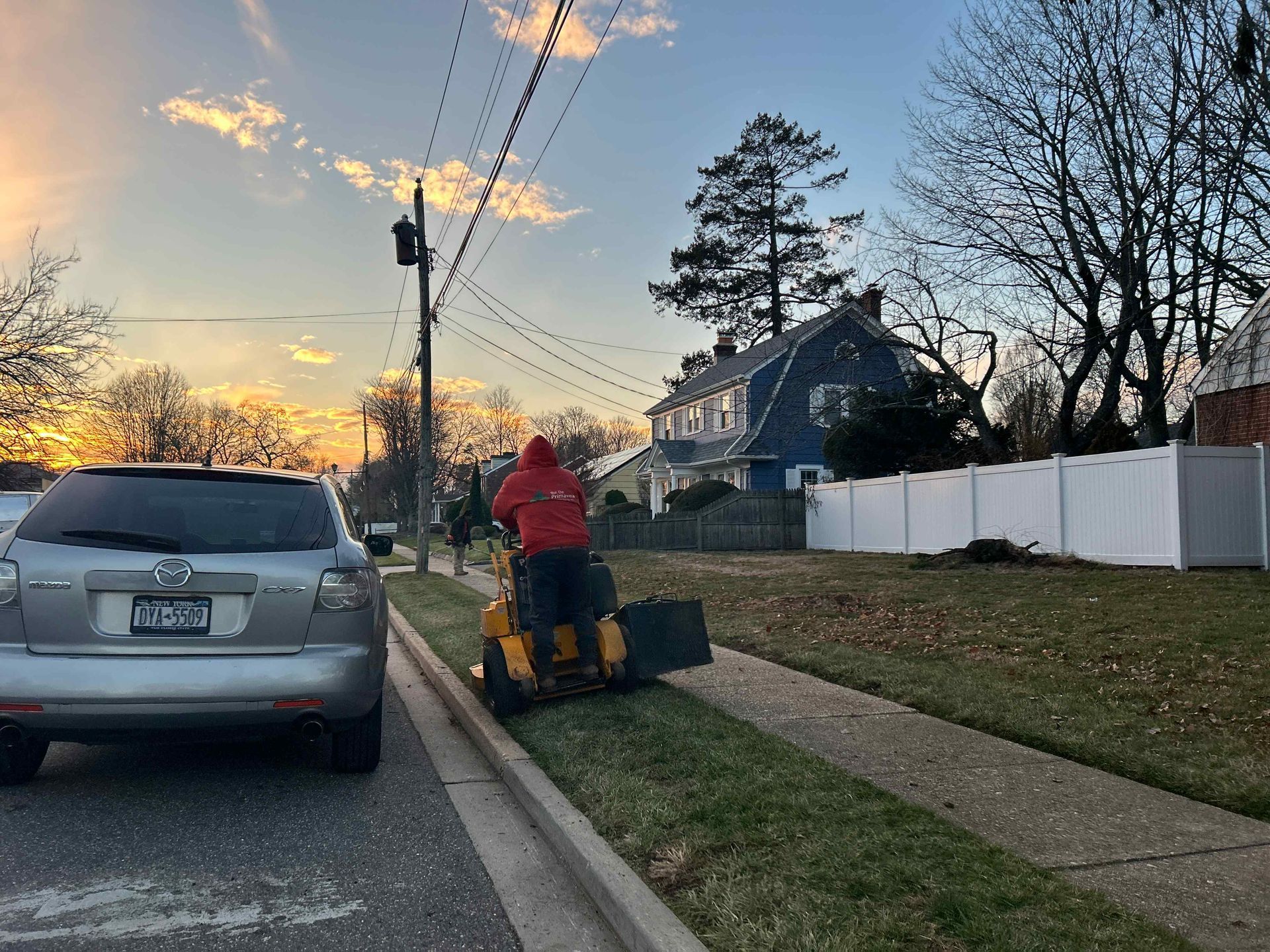 Person mowing a grassy roadside strip beside parked cars at sunset, with houses and trees in the background.