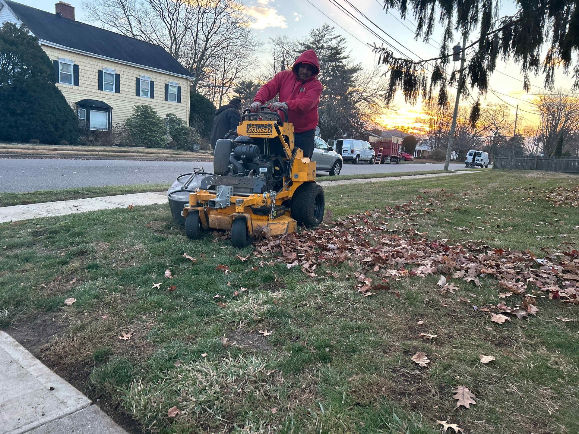 Person in red jacket using a yellow mower to clear fallen leaves on a lawn at sunset