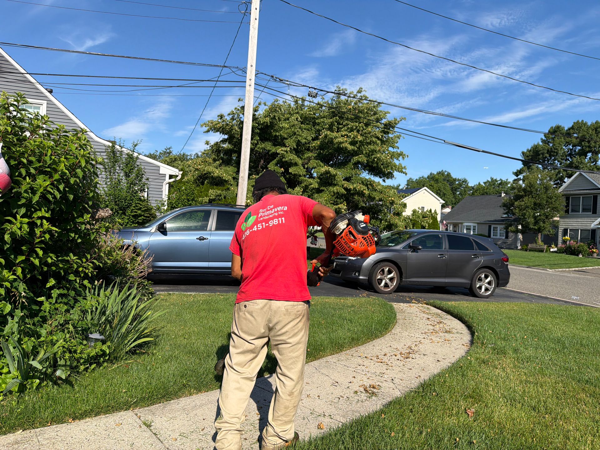 Person in red shirt on a lawn path near parked cars in a suburban neighborhood under a blue sky