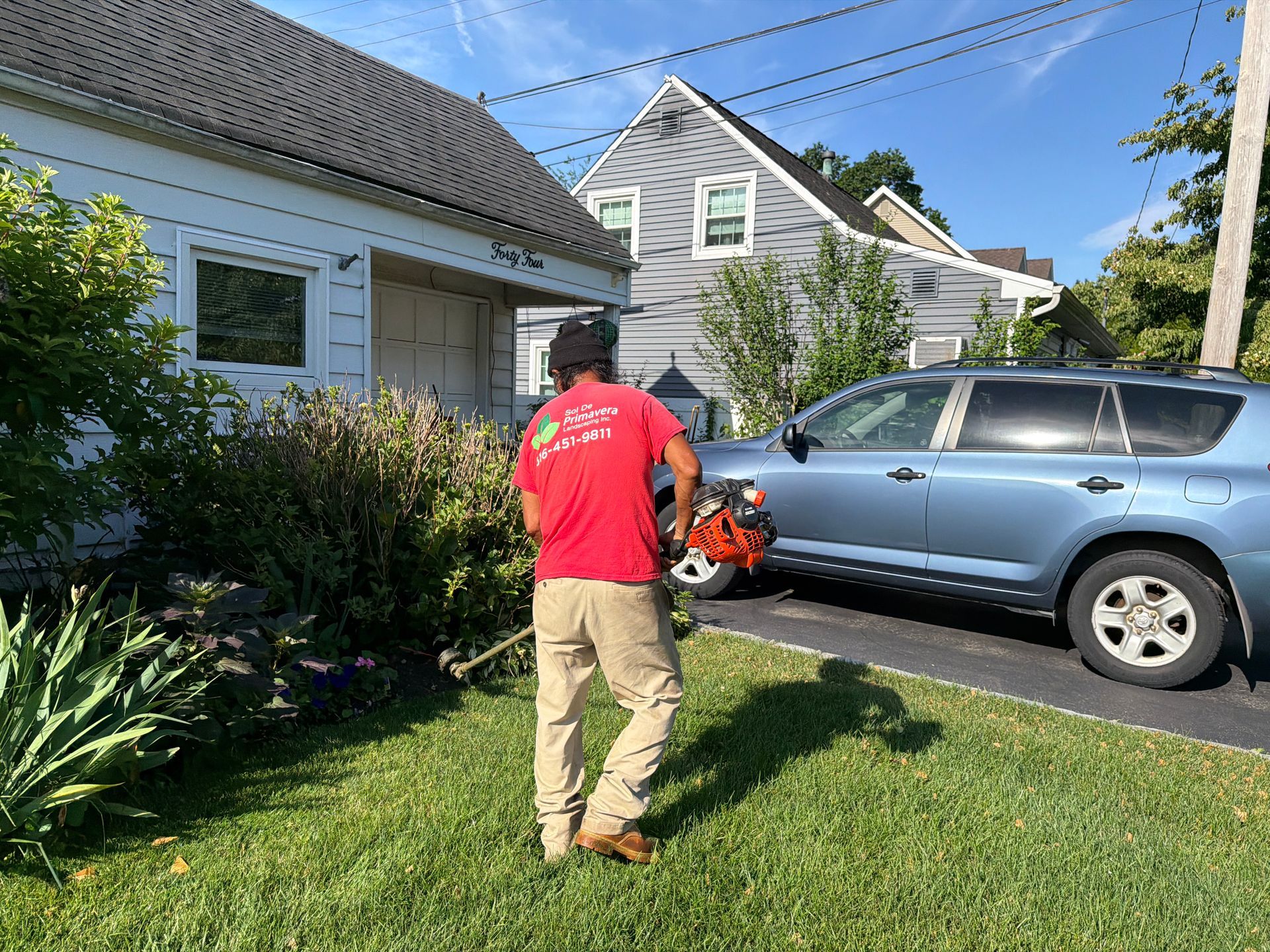 Person in red shirt using a leaf blower on a driveway beside a silver SUV and house.