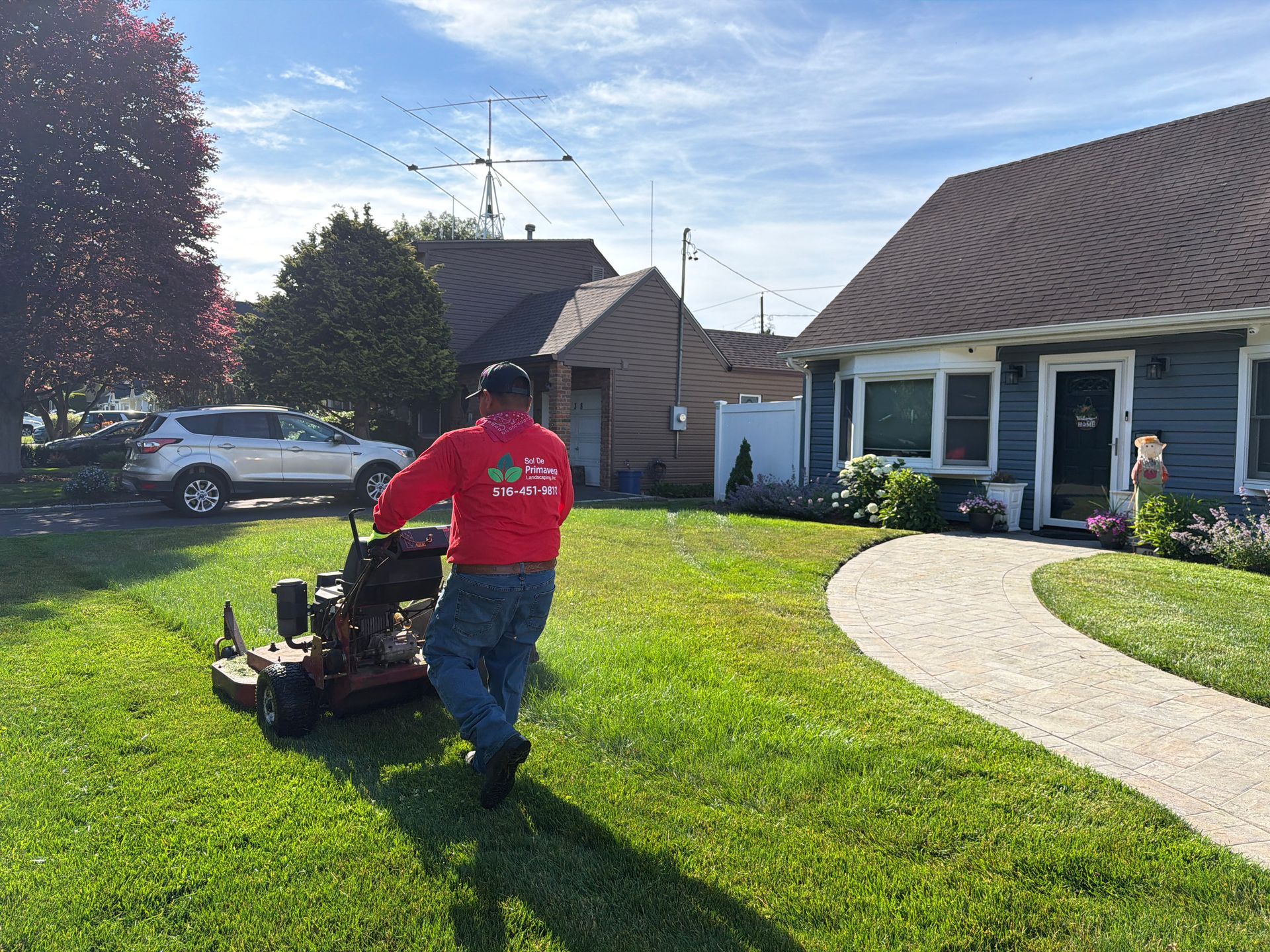Person in red shirt mowing a lawn beside a suburban house on a sunny day