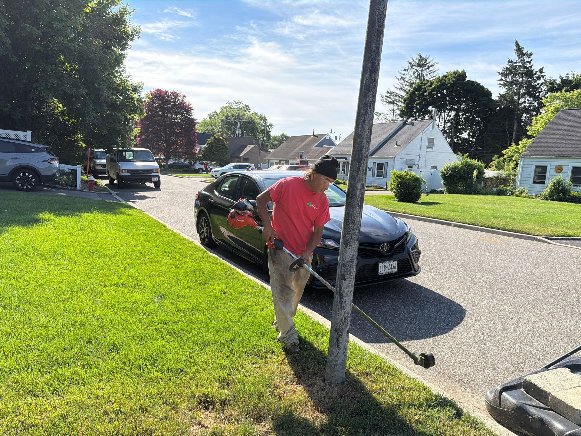 Person in pink shirt leaning on a pole beside a black car on a sunny suburban street.