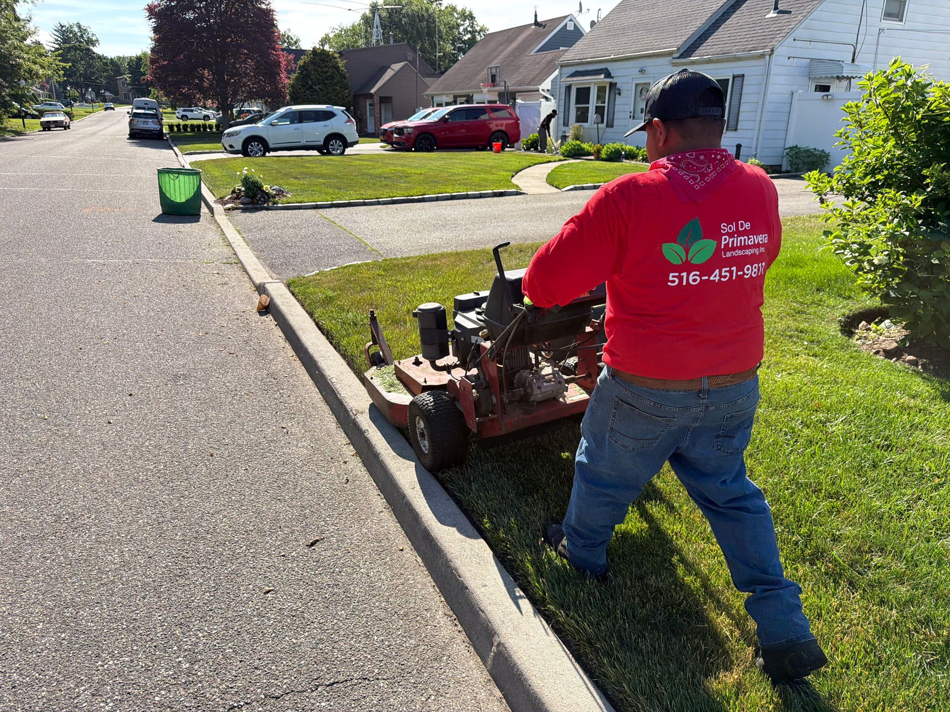 Worker in red shirt mowing a grassy curb strip in a suburban neighborhood.
