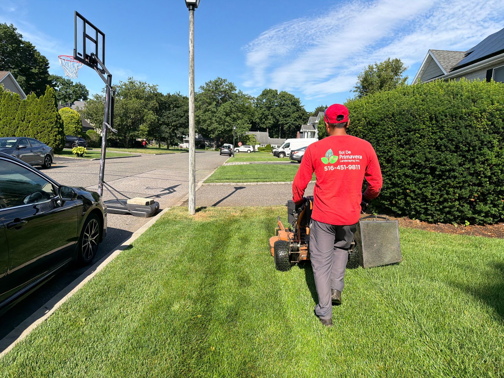 Person in red shirt mowing a lawn beside a driveway on a sunny suburban street.