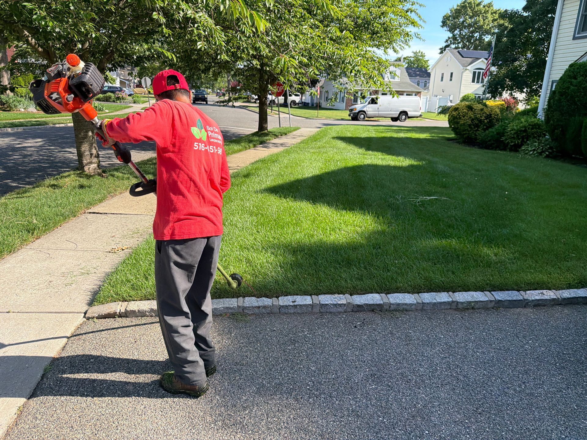 Worker in red shirt using a leaf blower along a sidewalk beside a grassy yard.