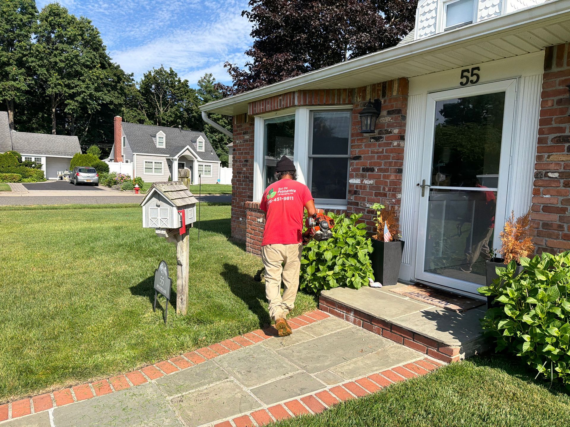 Person in a red shirt watering plants beside a brick house with an open front door.