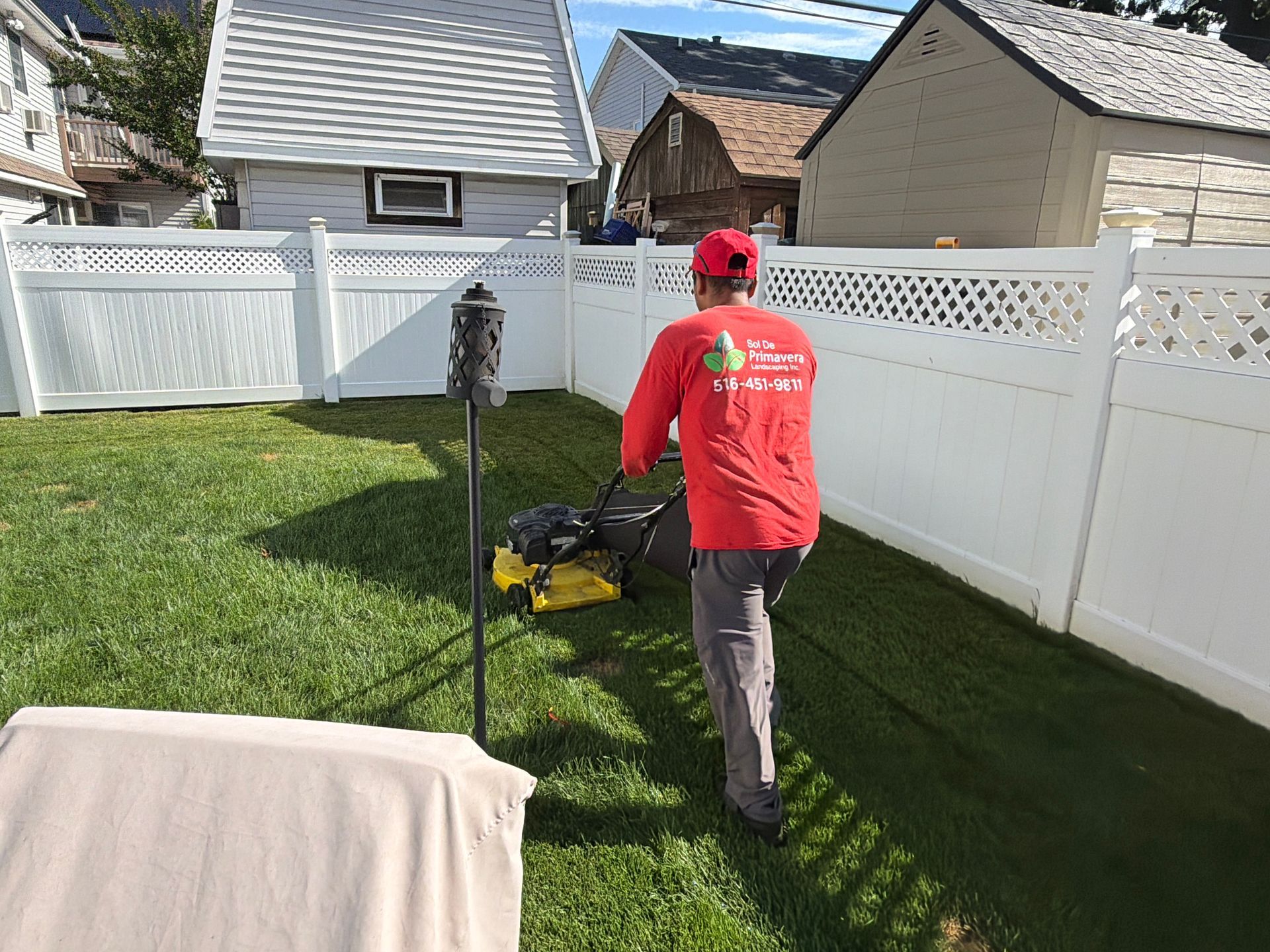 Person mowing a narrow strip of grass beside a white fence in a backyard