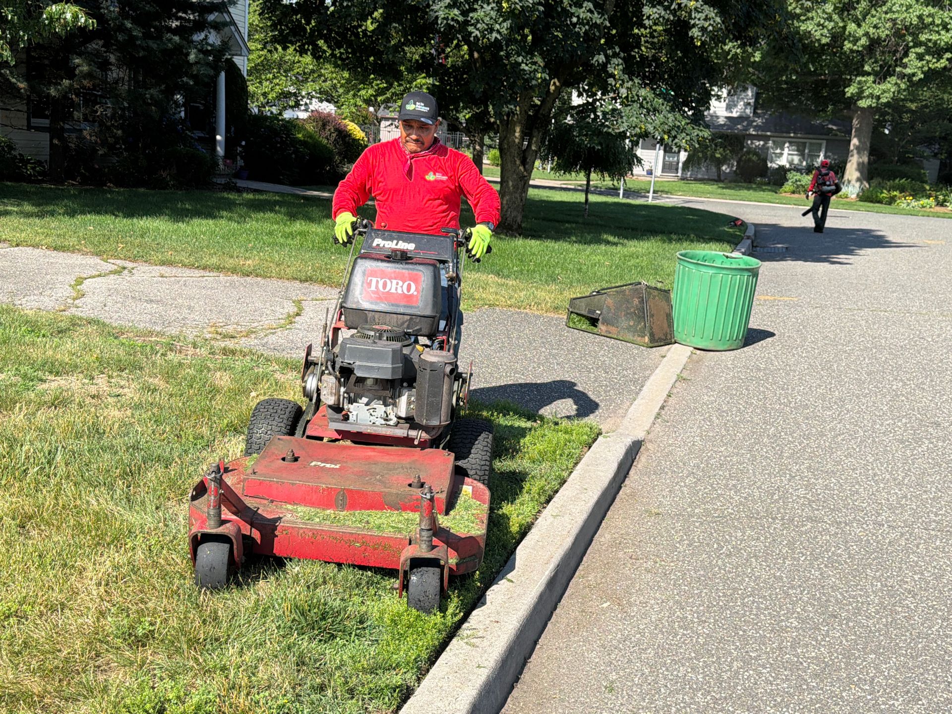 Person mowing a grassy roadside with a red riding mower beside a green trash bin