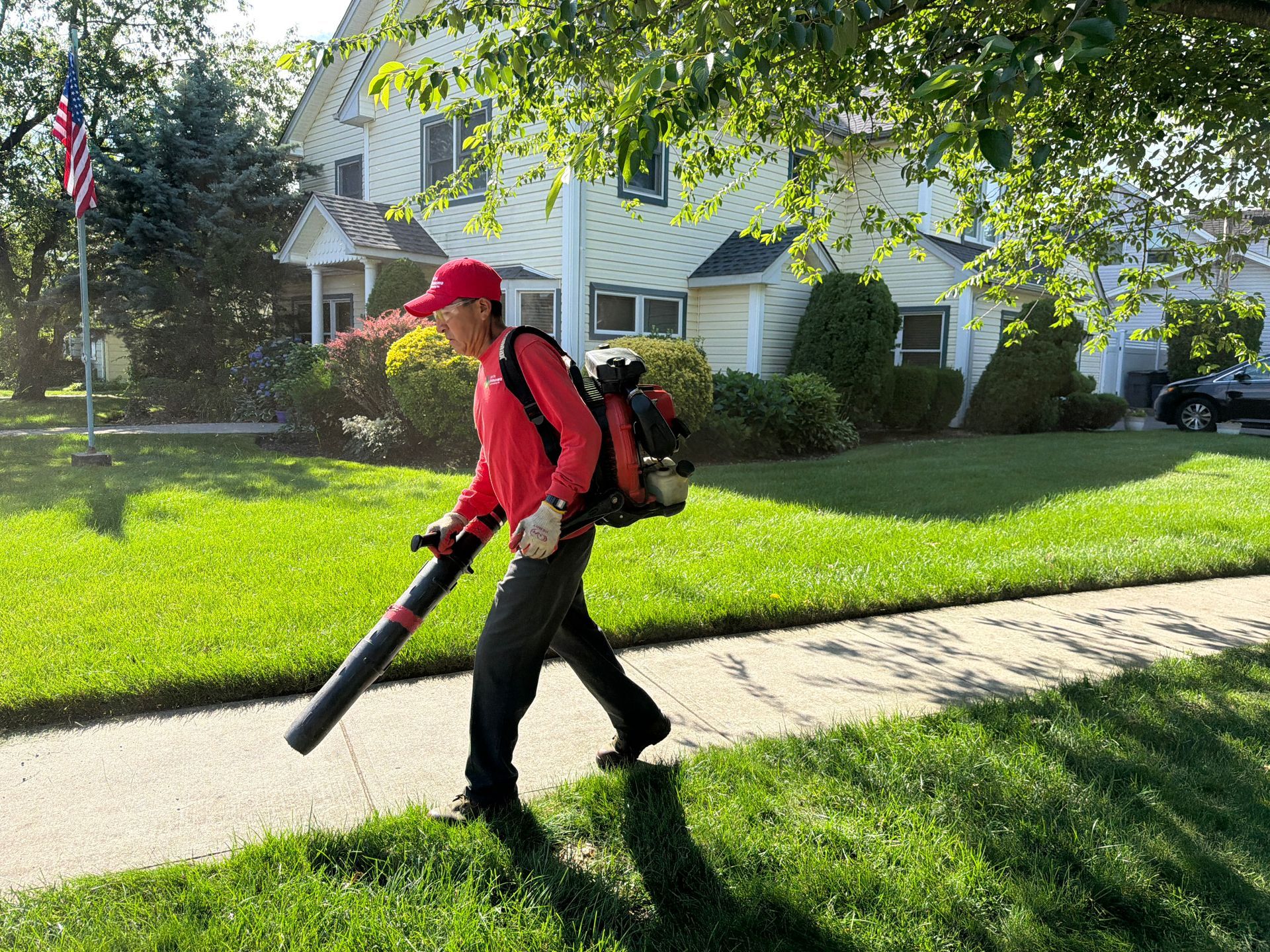 Person in red shirt using a leaf blower on a sunny suburban sidewalk.